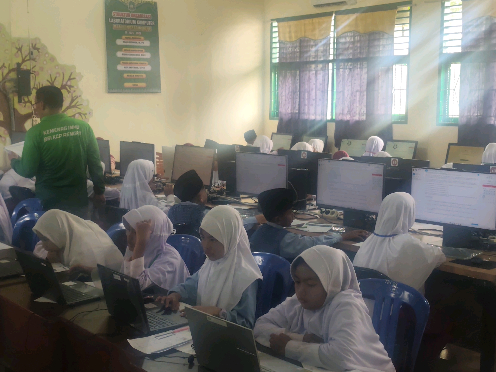The image depicts an indoor scene, likely a computer laboratory, with several young individuals, mostly wearing white hijabs, seated at desks with laptops and computer monitors. A man in a green shirt, identified by text as "KEMENAG INHU BSI KCP RENGAT," stands on the left, seemingly instructing or observing. The room is brightly lit by sunlight streaming through windows covered by yellow and purple curtains. A bulletin board with organizational structure information is visible on the wall. The presence of computer equipment, desks, and chairs suggests an educational setting. The text on the man's shirt and the overall environment point to the location being a computer laboratory associated with Kemenag Inhu, BSI KCP Rengat, likely in or near Pematang Reba, Indonesia.