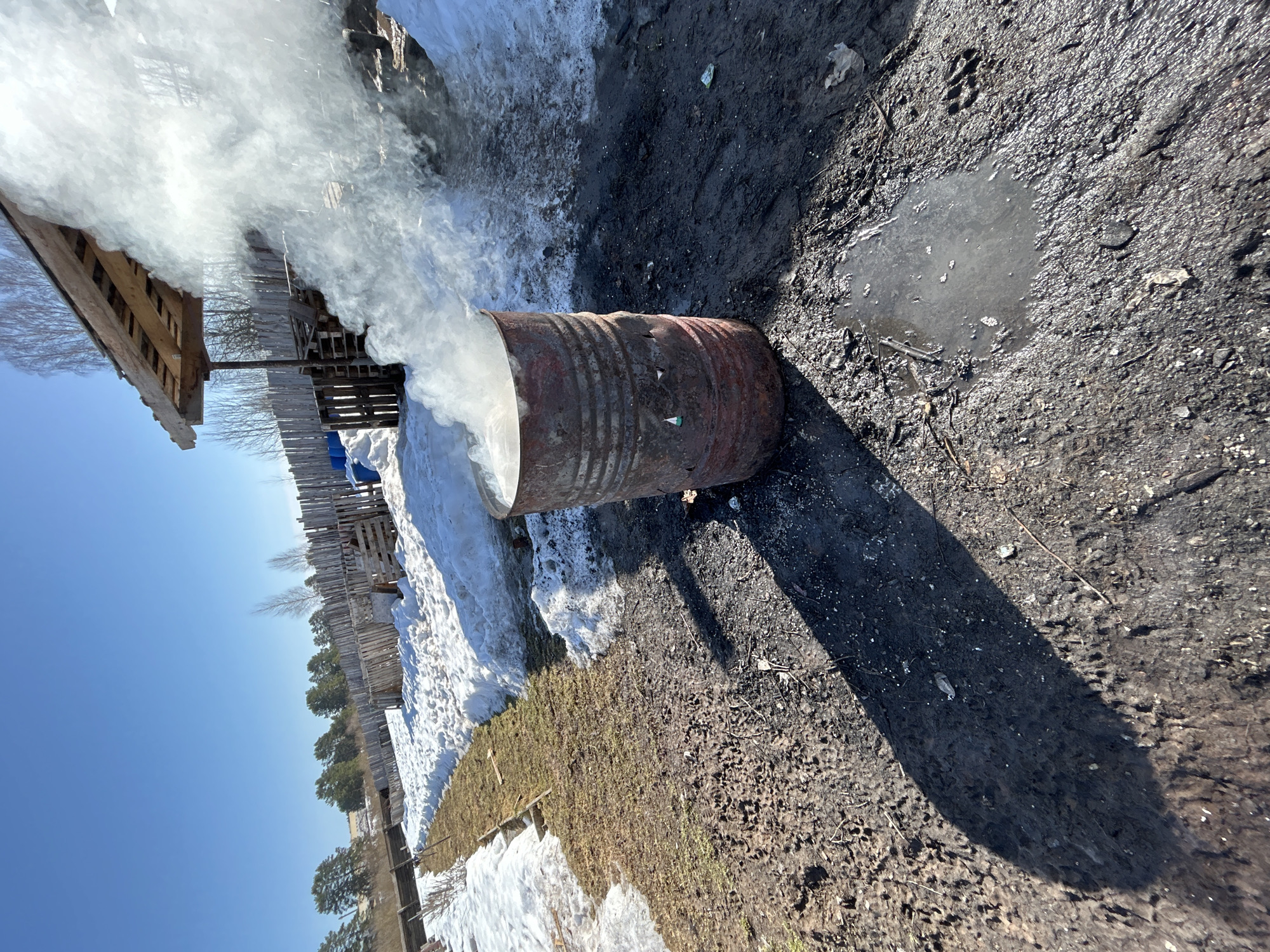 The image shows a metal barrel lying horizontally on the ground with a large plume of white smoke emanating from its open end. The barrel appears rusty and weathered. The ground is a mix of dirt, mud, and some snow. A small puddle of water is visible near the right edge of the image. In the background, there is a wooden fence and a shed-like structure. The sky is blue and clear. Based on the presence of snow and the general appearance of the environment, the location is likely in a rural or semi-rural area, possibly near Rusalevka, Russia. There are no people visible in the image. The scene suggests the burning of some material within the barrel.