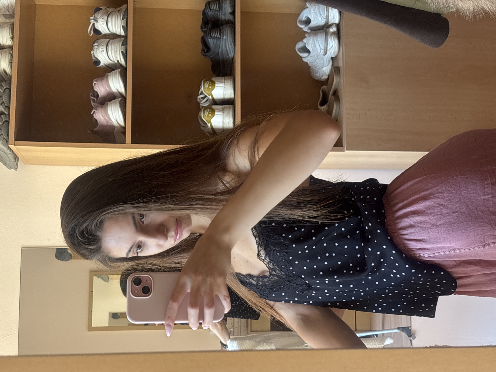 The media file shows a young woman taking a mirror selfie in what appears to be a closet or dressing area. The woman has long, dark brown hair and is wearing a black top with white polka dots and a pink skirt. She is holding a pink smartphone to take the photo.
The background reveals shelves filled with various shoes, including sneakers and what look like casual slides. The shelves are made of light-colored wood. In the reflection of the mirror, another smaller mirror is visible, as is a portion of the room, which seems to have light-colored walls.
The lighting suggests it is daytime, possibly indoors with artificial or natural light. The overall impression is casual and personal, capturing a moment of self-documentation. There is no visible text in the image. The implied location is Varna, Bulgaria, as stated.