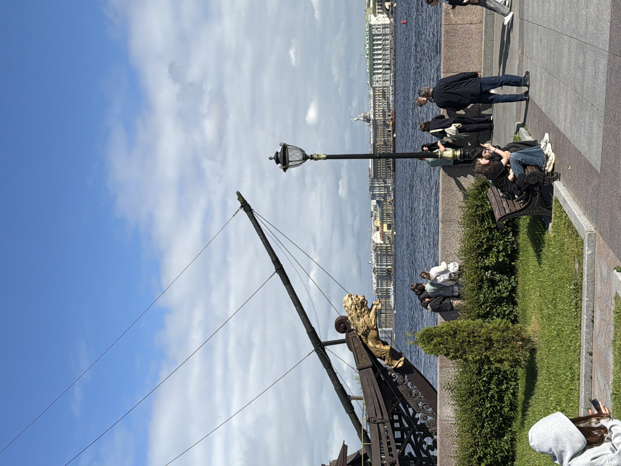 The image depicts a daytime scene on a waterfront promenade in Saint Petersburg, Russia. In the foreground, on the left, a person wearing a grey hooded sweatshirt is sitting down and looking at a mobile phone. To the right, a couple sits on a park bench, with one person pointing outwards. Further along the promenade, a group of people are standing and conversing near a hedgerow, with a tall lamppost to their right. In the background, a large body of water, presumably the Neva River, separates the promenade from the city's architectural landmarks, including a long row of classical buildings and a dome. Dominating the left side of the image is the bow of a decorative ship, featuring intricate dark wood carvings and a golden lion's head figurehead. The sky is blue with scattered white clouds. The overall atmosphere suggests a pleasant day for a stroll in the city.