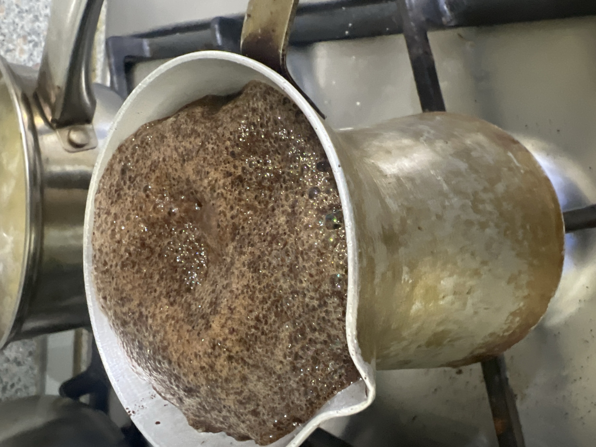 The image captures a close-up, top-down perspective of coffee actively brewing in a metal Cezve (Turkish coffee pot) on a stovetop. The coffee inside the Cezve is dark brown and covered with a thick, frothy, bubbling foam, indicating it is heating up. Small, shimmering highlights are visible on the surface of the foam.
The Cezve itself appears to be made of metal, possibly copper or brass, and shows signs of wear or patina. It has a single, straight handle that extends upwards. The pot is positioned over a dark-colored gas burner grate, suggesting a gas stovetop is in use.
To the left of the Cezve, a portion of another metallic pot with a similar handle is visible, resting on a light-colored kitchen surface with a mottled or granite-like pattern. The background further shows indistinct elements of a kitchen environment, including more of the stovetop and possibly a wall.
The activity depicted is the preparation of coffee, likely Turkish-style, in a home kitchen. There are no people visible in the image, nor any discernible text. The scene conveys a simple, domestic moment of beverage preparation. The location context of Chaikovsky, Russia, is consistent with the type of coffee pot shown. Time of day and weather cannot be determined from the indoor setting.