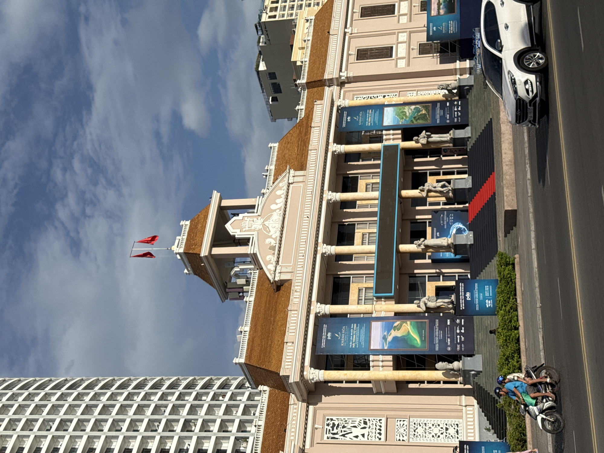 The image shows a large, ornate building with a pink facade and a brown tiled roof, located in Nha Trang, Vietnam. The building features a classical architectural style with columns and decorative elements. Two Vietnamese flags are visible on the roof. In front of the building, banners displaying scenic views and text in Vietnamese are hung. A red carpet leads up a set of stairs to the building's entrance, flanked by statues. To the left of the building, a tall, modern skyscraper with a grid-like pattern of windows is visible. In the foreground, a white SUV is driving on a street, and a motorcycle with two riders is also on the street. The scene appears to be set during the daytime, with clear skies and shadows indicating the sun is at an angle. The overall impression is of a grand public building in a city setting, possibly a cultural or governmental institution, with contemporary urban elements surrounding it.