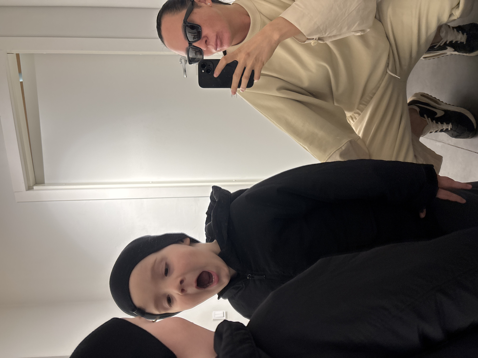 A selfie-style photograph taken indoors shows a woman in sunglasses holding a smartphone and kneeling to take a picture. She is wearing a light beige outfit and black and white sneakers. A child wearing a black beanie and a black jacket is in the foreground, appearing to look upwards at the camera. The location appears to be a dressing room, indicated by the walls and door frame in the background. The city of Chaikovsky, Russia, cannot be confirmed from the given image.