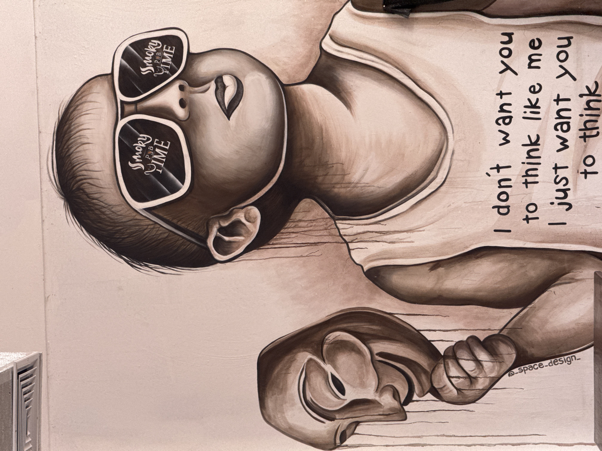 This is a mural painted on a wall, likely in Higüey, Dominican Republic, depicting a stylized infant wearing sunglasses. The sunglasses have "Smoky PUB TIME" written on them, with the words separated by a graphic of a pipe. The infant is shown with its head tilted back, eyes closed, and a peaceful expression. The mural is rendered in shades of brown and beige, giving it a monochromatic and artistic feel.
The infant is wearing a simple tank top. In the infant's hand, it is holding a Guy Fawkes mask. To the right of the infant, text is written vertically on the wall. The text reads: "I don't want you to think like me I just want you to think". Below this text, a signature or artist's mark is visible: "@_space.design_".
The style of the artwork is illustrative and somewhat surreal, blending elements of innocence (the infant) with a message of individuality and critical thinking (the text and the mask, often associated with anonymity and rebellion). The lighting and shadows suggest an indoor setting or a shaded outdoor area, and there are no clear indicators of the time of day or weather. The overall impression is one of contemplation and a call to independent thought. A ventilation grille is visible in the bottom left corner, suggesting the mural is on an interior wall or an exterior wall adjacent to a building.