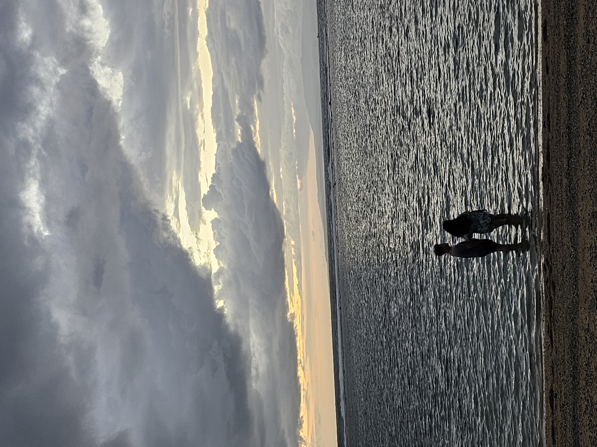 Two individuals stand in shallow water at the edge of a beach, facing away from the camera and towards the ocean. The person on the left is wearing dark shorts and a t-shirt, while the person on the right is wearing a patterned dress. The water is dark and rippled, with a faint line of breaking waves visible further out. In the background, a distant landmass, likely islands, is silhouetted against a dramatic sky filled with dark, heavy clouds interspersed with patches of lighter sky and hints of yellow and orange light near the horizon. The scene is set at dusk on a beach in Denpasar, Indonesia, with the ocean and a cloudy sky dominating the composition.