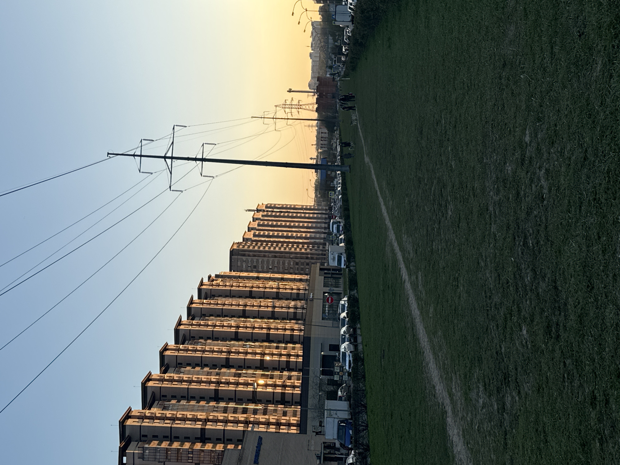 The image shows a view in Odintsovo, Russia, at sunset. A line of residential buildings is prominently featured, their facade illuminated by the low sun. To the right of the buildings, there is a grassy field with a faint white line marking a boundary. A row of parked cars separates the buildings from the field. Overhead power lines are visible against the sky. In the distance, additional buildings and street lights can be observed, creating an urban backdrop.