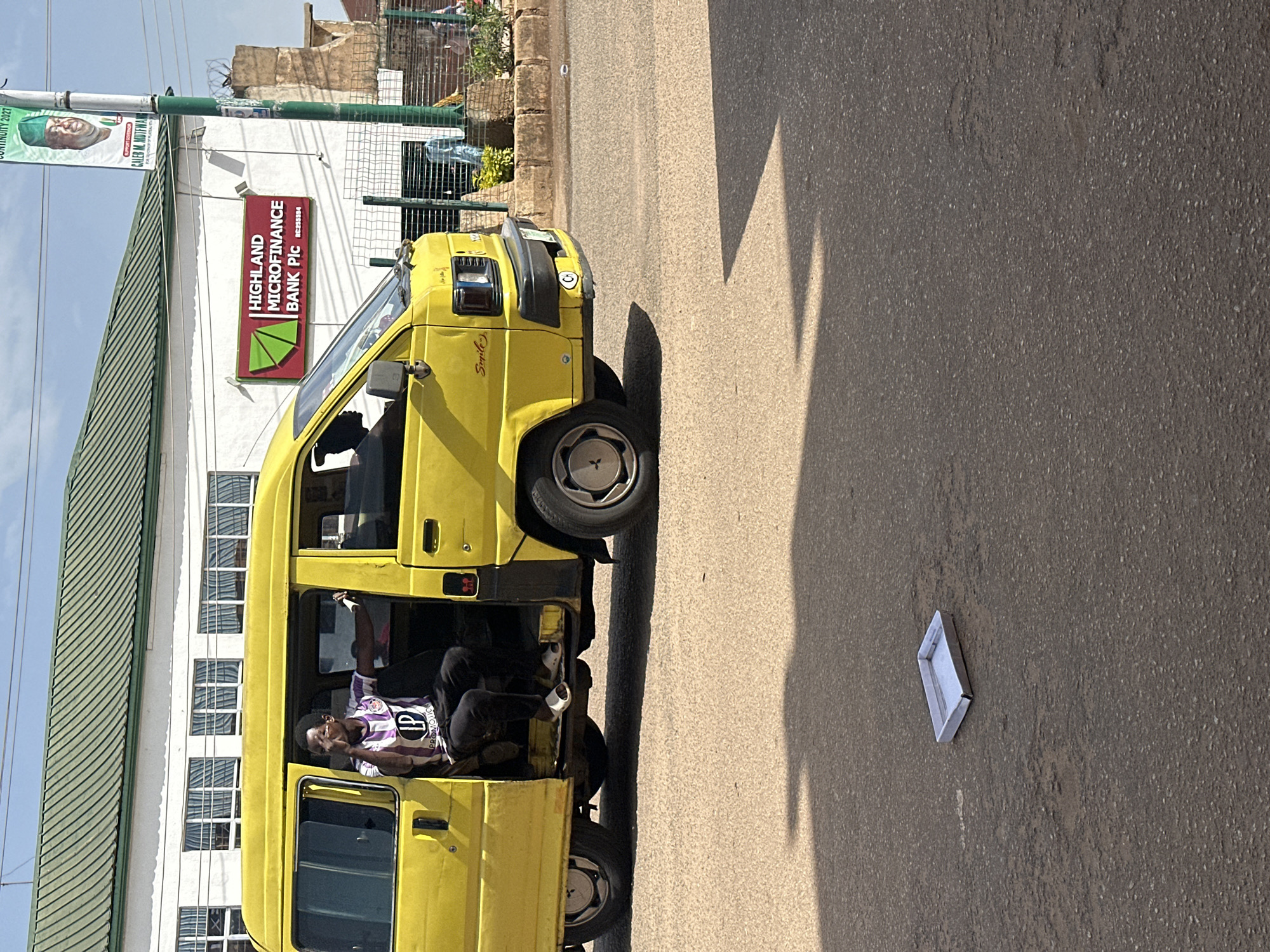 In Bukuru, Nigeria, a bright yellow minivan is parked on a paved road. The driver's side door is open, and a young man wearing a purple and white striped shirt with the logo "LP Promotion" and black pants is seated inside, looking towards the right of the frame. He is wearing white slippers. The minivan has "Smile" written on its front. Behind the minivan, a white building with a green roof has a prominent red sign that reads "HIGHLAND MICROFINANCE BANK PLC." A green banner displaying a portrait of a man and text related to "CONTINUITY 2027" hangs from a pole to the right of the bank. In the foreground, a small, empty metal tray rests on the asphalt. The road is marked with shadows cast by unseen objects.