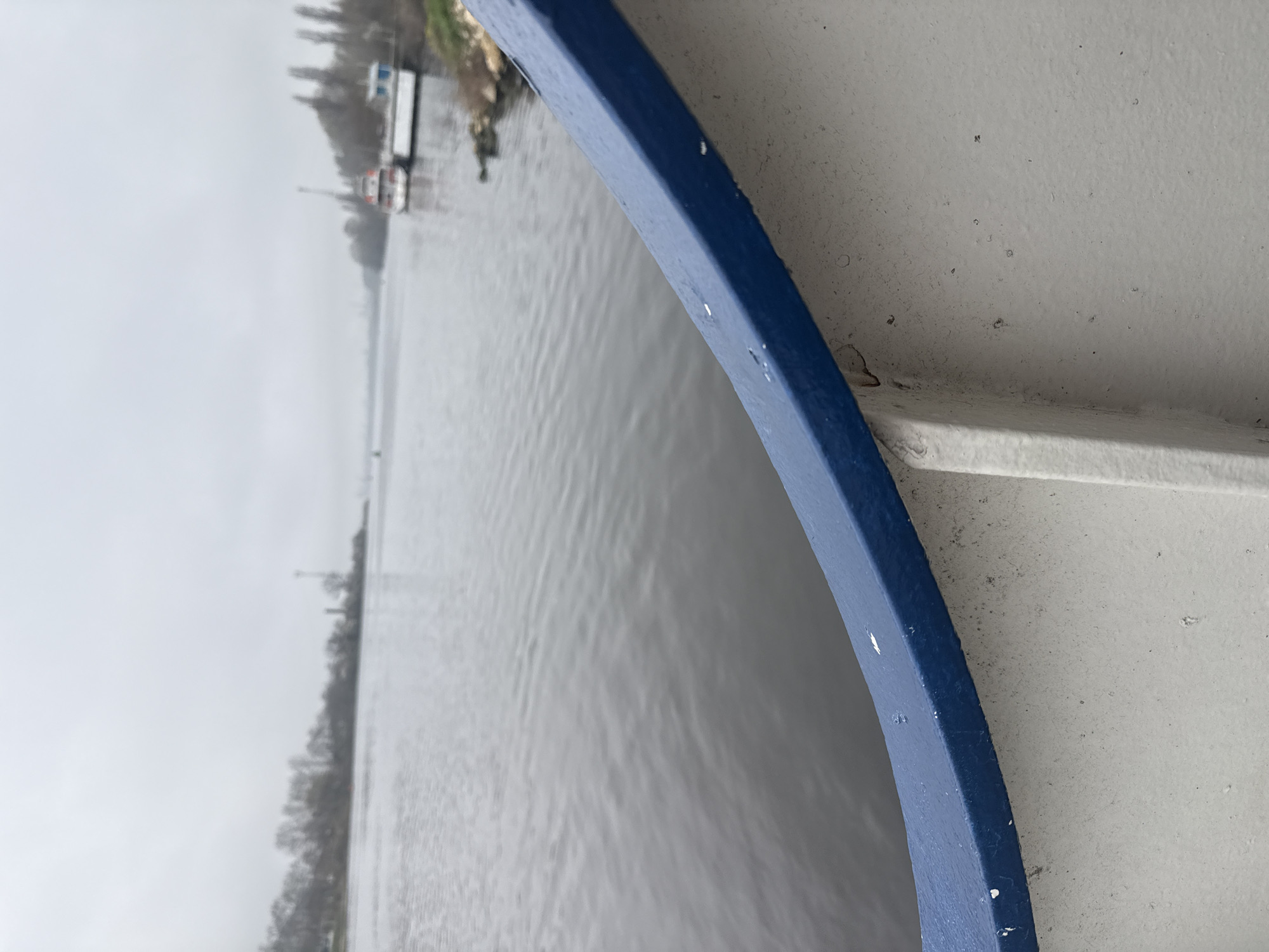 The image, taken from a vessel on a waterway, shows a river scene on an overcast day. A blue railing is in the foreground. Across the water, there is evidence of land with trees. Further in the distance, there appear to be buildings or docked boats. The scene is likely near Beloslav, Bulgaria, as indicated in the prompt. There are no people visible in the frame. The water's surface is somewhat choppy.