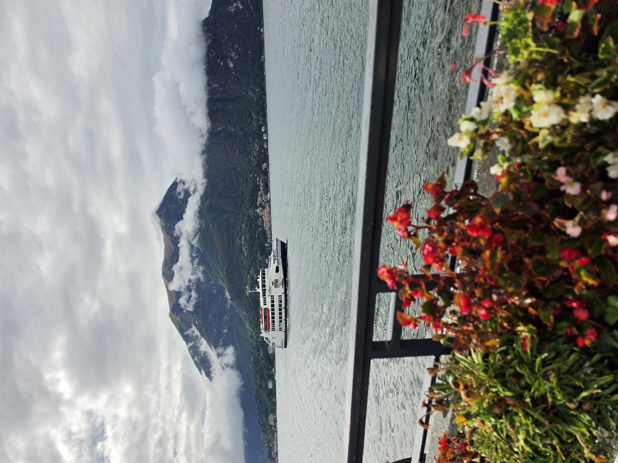 A white ferry boat travels across a gray, choppy lake. In the background, a green, mountainous landscape is partially obscured by swirling gray clouds. In the foreground, a railing separates the viewer from the lake, and a planter filled with red and white flowers is in focus. The scene is outdoors, with overcast skies suggesting a cool or humid day. The location is San Giovanni, Italy.