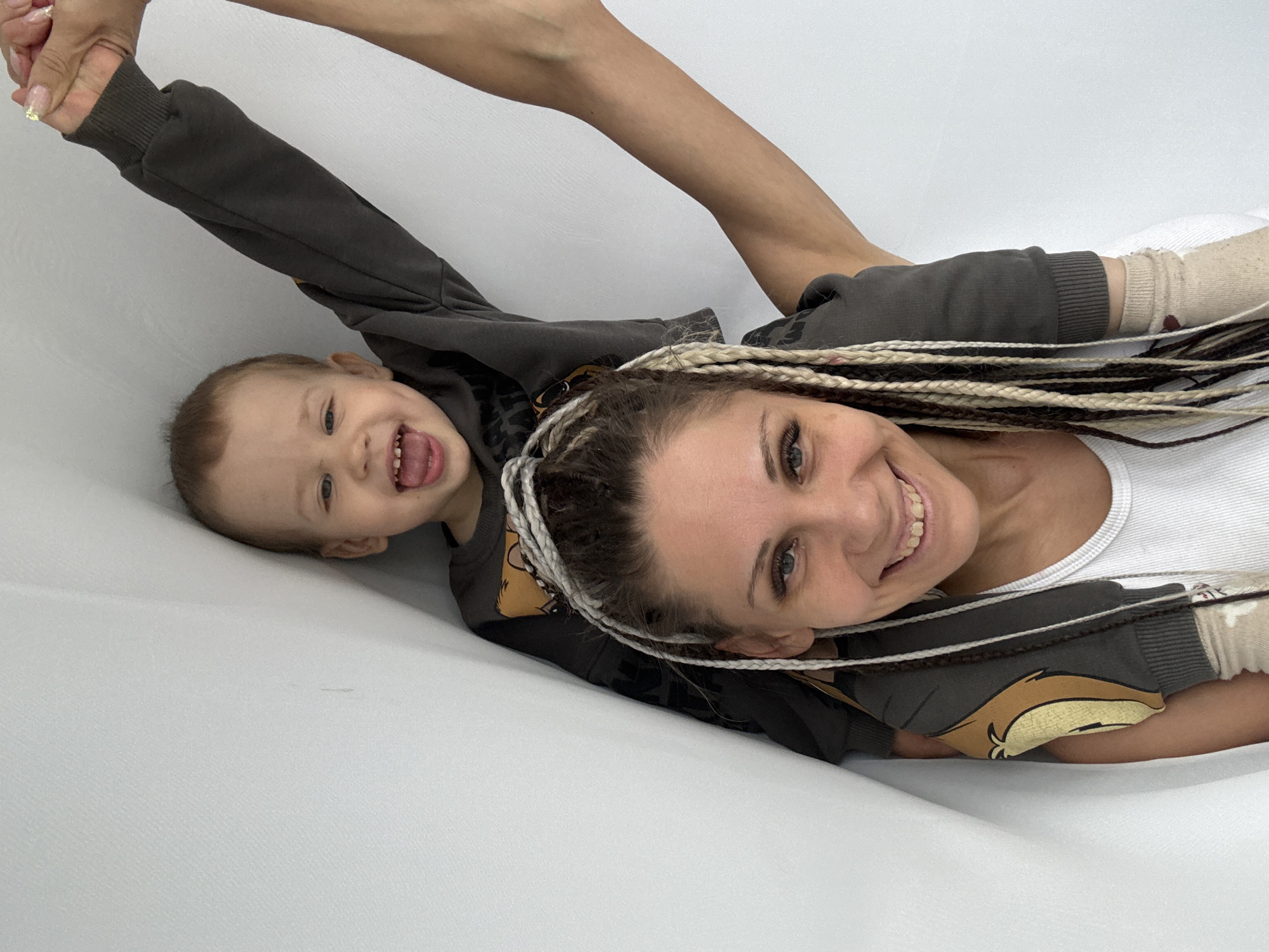 This vibrant portrait captures a joyful moment between a woman and a young child, possibly in Rusanovo, Russia. Both are lying down on a plain white surface, looking up at the camera with broad smiles.
The woman, with a distinctive hairstyle of blonde and brown dreadlocks, wears a white tank top and a dark gray hooded sweatshirt. Her blue-green eyes sparkle as she smiles, revealing her teeth.
Beside her, a young child, likely a toddler, with light brown hair and blue eyes, is also clad in a similar dark gray hooded sweatshirt, which features a cartoon-like animal face, possibly a squirrel or chipmunk, on the front. The child is laughing exuberantly, with an open mouth and tongue visible, conveying pure delight.
An adult's arm and hand, showing manicured nails, extend from the top of the frame, gently holding the child's small hand, suggesting a playful interaction or a shared moment of connection. The overall mood of the image is one of happiness, affection, and lighthearted fun. The pristine white background serves to highlight the subjects and their joyful expressions, making them the central focus of the scene.