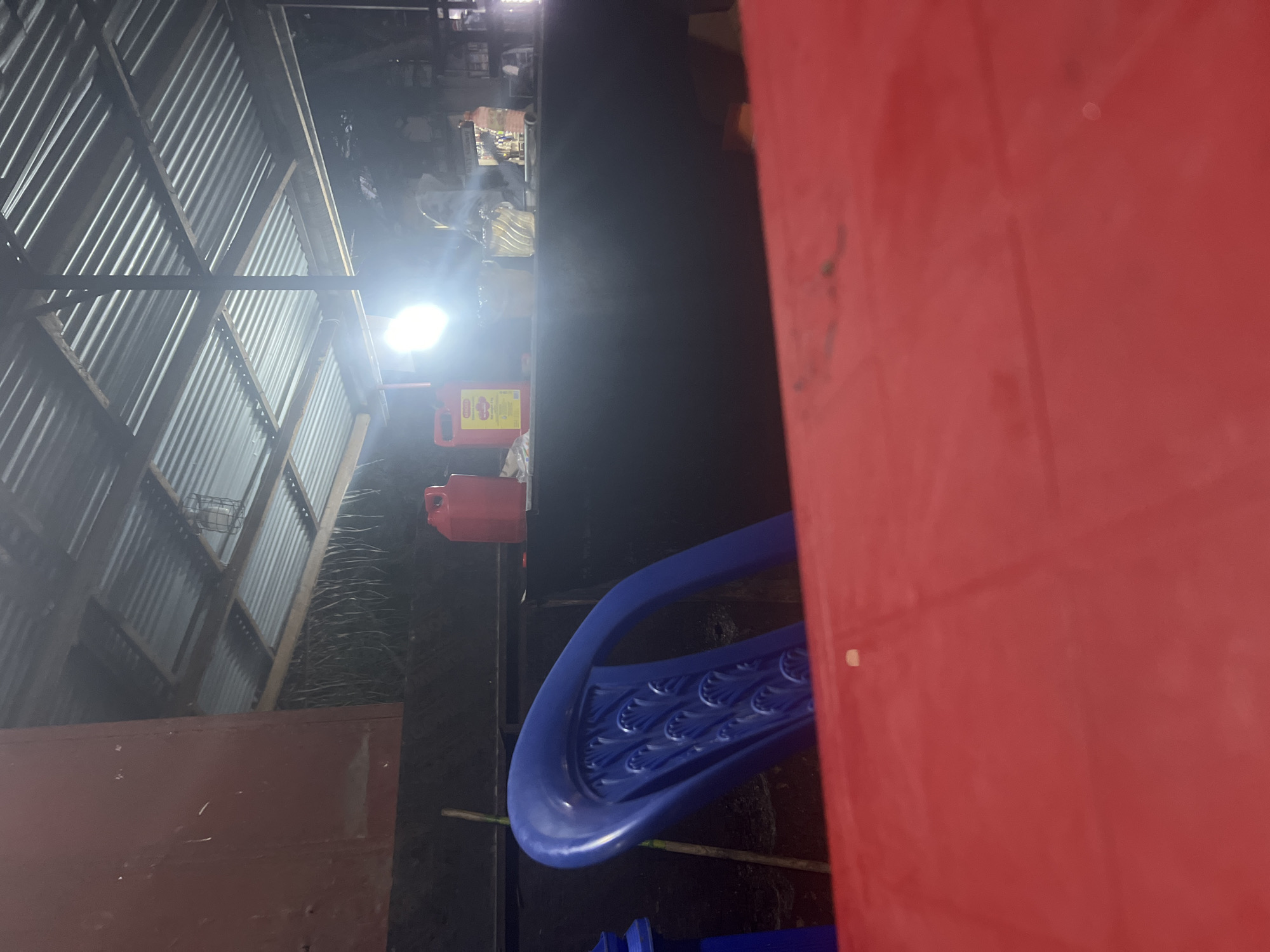 The image shows a partially visible red structure, possibly a building or large object, dominating the right side of the frame. A blue structure with patterned detailing is positioned near the bottom. In the background, there is a corrugated metal structure that appears to be part of a building's roof or wall. A bright light source illuminates the scene, casting shadows. There are various unidentified objects and materials scattered around the area. The location cannot be determined based on the visual information provided.