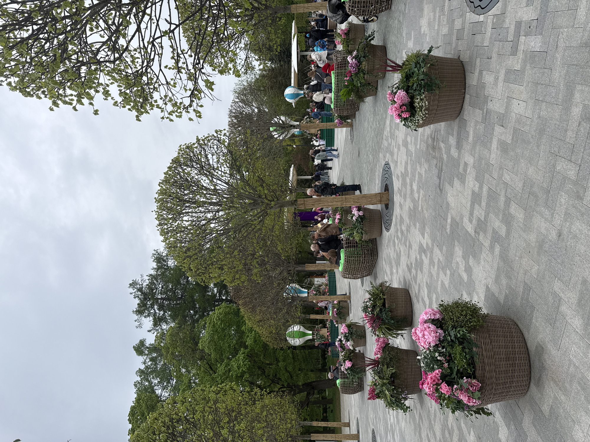 The image shows a public space in Moscow, Russia. The area is paved with gray stone in a geometric pattern. Along one side of the paved area are trees planted in the ground with woven material wrapped around their trunks. Large woven baskets filled with pink flowers and green foliage are placed near the base of the trees. In the background, there are more trees and shrubs, as well as some people. There are also decorations resembling hot air balloons. Benches are situated along the walkway and are occupied by individuals. The sky is overcast.