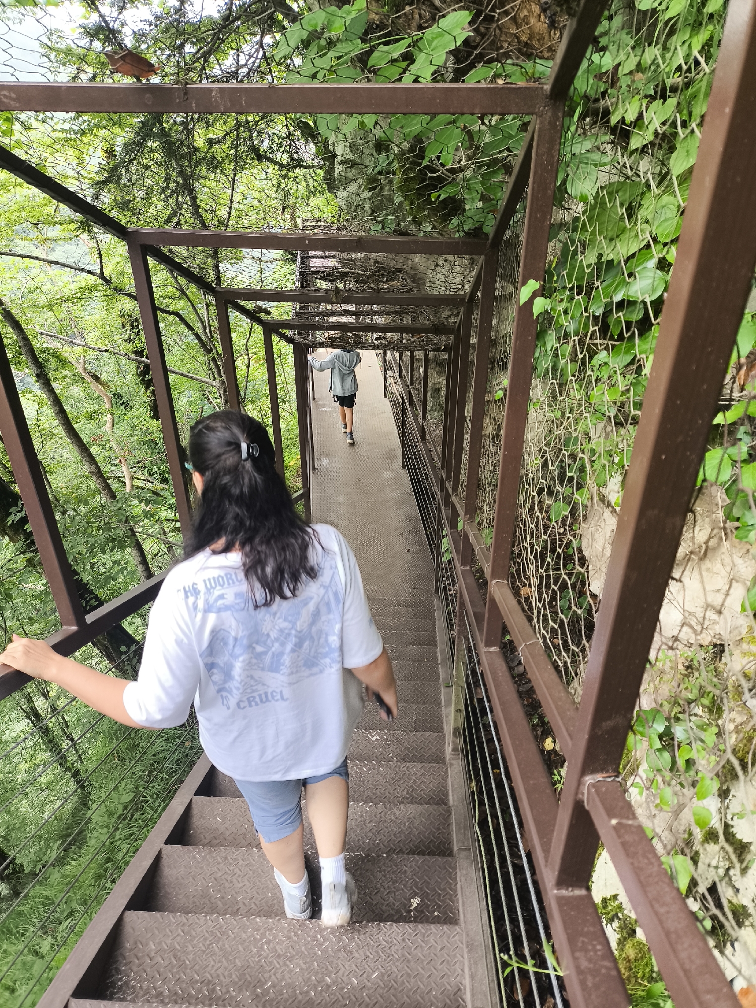 In the city of Satsiskvilo, Georgia, a metal staircase with brown railings descends through a lush, green, mountainous area. The stairs are made of diamond plate metal, and the railings are a dark brown color. The staircase is bordered on one side by a wire mesh fence, behind which dense foliage and trees are visible. On the other side, the embankment is covered with greenery and some rock formations.
In the foreground, a woman with long, dark hair pulled back with a clip is walking down the stairs. She is wearing a white t-shirt with a graphic print on the back and light blue shorts. She is holding onto the railing with her left hand and appears to be holding a mobile phone in her right hand.
Further down the staircase, another person, dressed in a grey hoodie and dark shorts, is walking away from the camera. The environment is characterized by dense vegetation and a slightly overcast sky, suggesting a natural, outdoor setting likely part of a tourist path or trail.