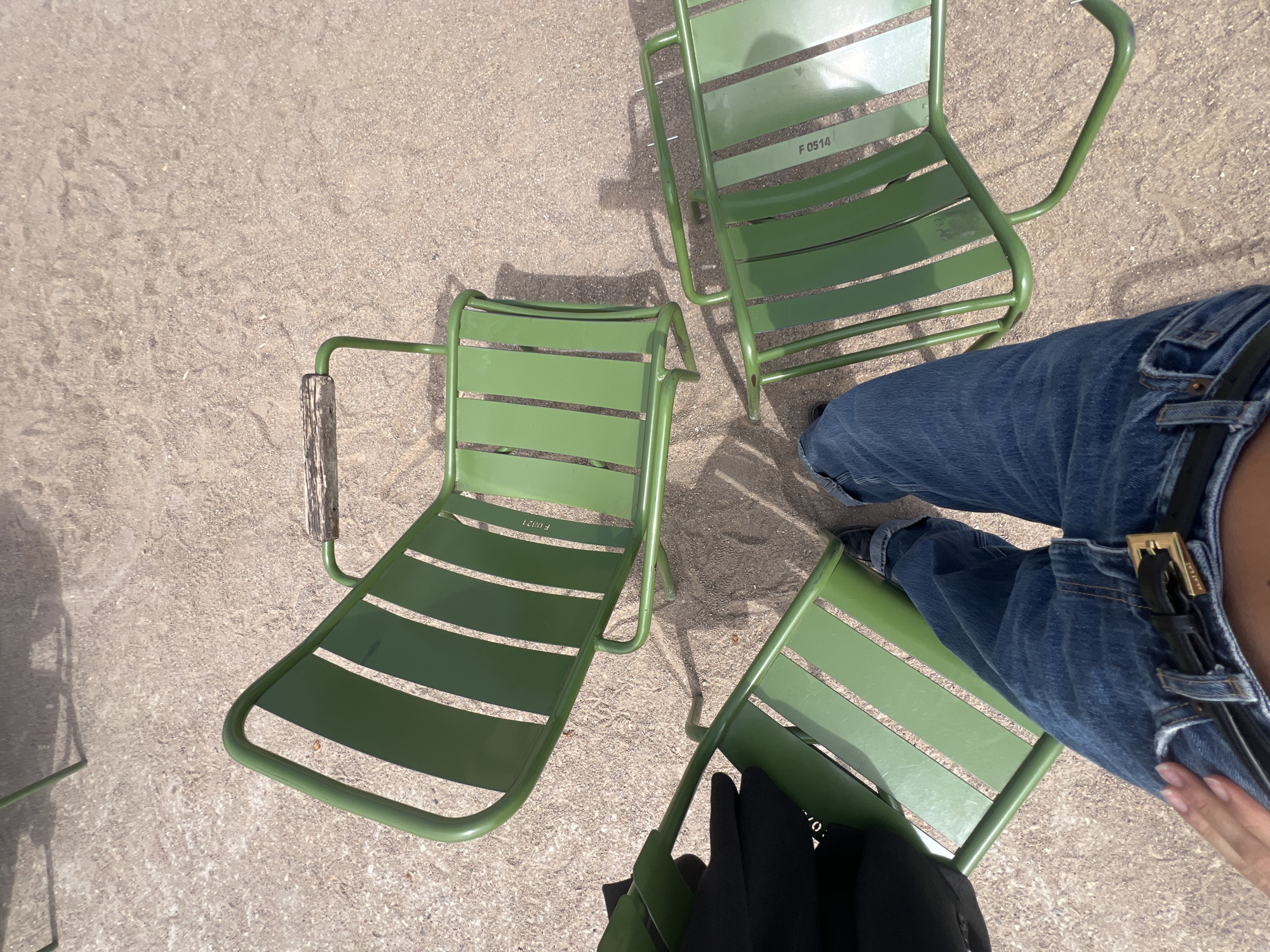 The image presents an overhead view of an outdoor setting, likely a park in Paris, France, under bright daylight. Three distinctive green metal chairs with slatted seats and backs are scattered on a light brown, gravelly or sandy ground. Strong sunlight illuminates the scene, casting clear shadows from the chairs and the person.
The lower half of a person is visible on the right side of the frame. They are wearing blue denim jeans, dark shoes, and a dark belt with a prominent gold-toned buckle. A hand with neatly manicured nails is visible near the person's hip. One of the person's legs is draped over a chair, indicating they are either seated, in the process of sitting down, or adjusting their posture.
Notable text is visible on the slats of two chairs: "F 0514" is seen on the chair in the upper right portion of the image, and "F 0821" is visible on the chair in the lower left. These are likely identification or inventory numbers. The scene depicts a casual moment in a public green space, characteristic of Parisian outdoor leisure.