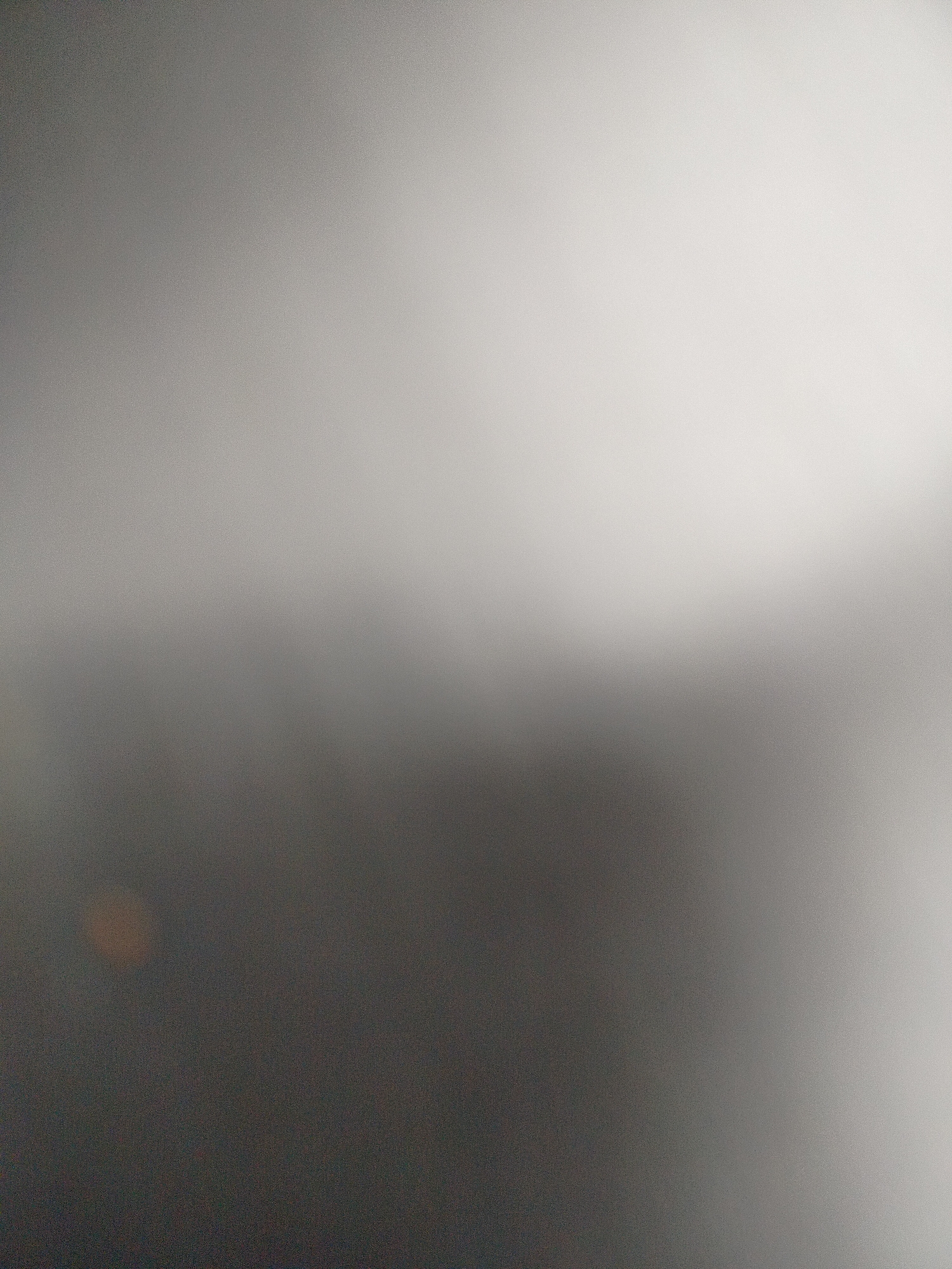 The image is a blurred, abstract gradient of gray and white. The lower portion is dark gray, gradually fading into a lighter gray, and then into a near-white hue at the top. There is a small, indistinct orange spot in the lower-left corner. The blurry quality makes it impossible to identify any specific objects or actions. There are no discernible people, subjects, or interactions depicted. The image does not contain enough information to locate it within Yerevan, Armenia.