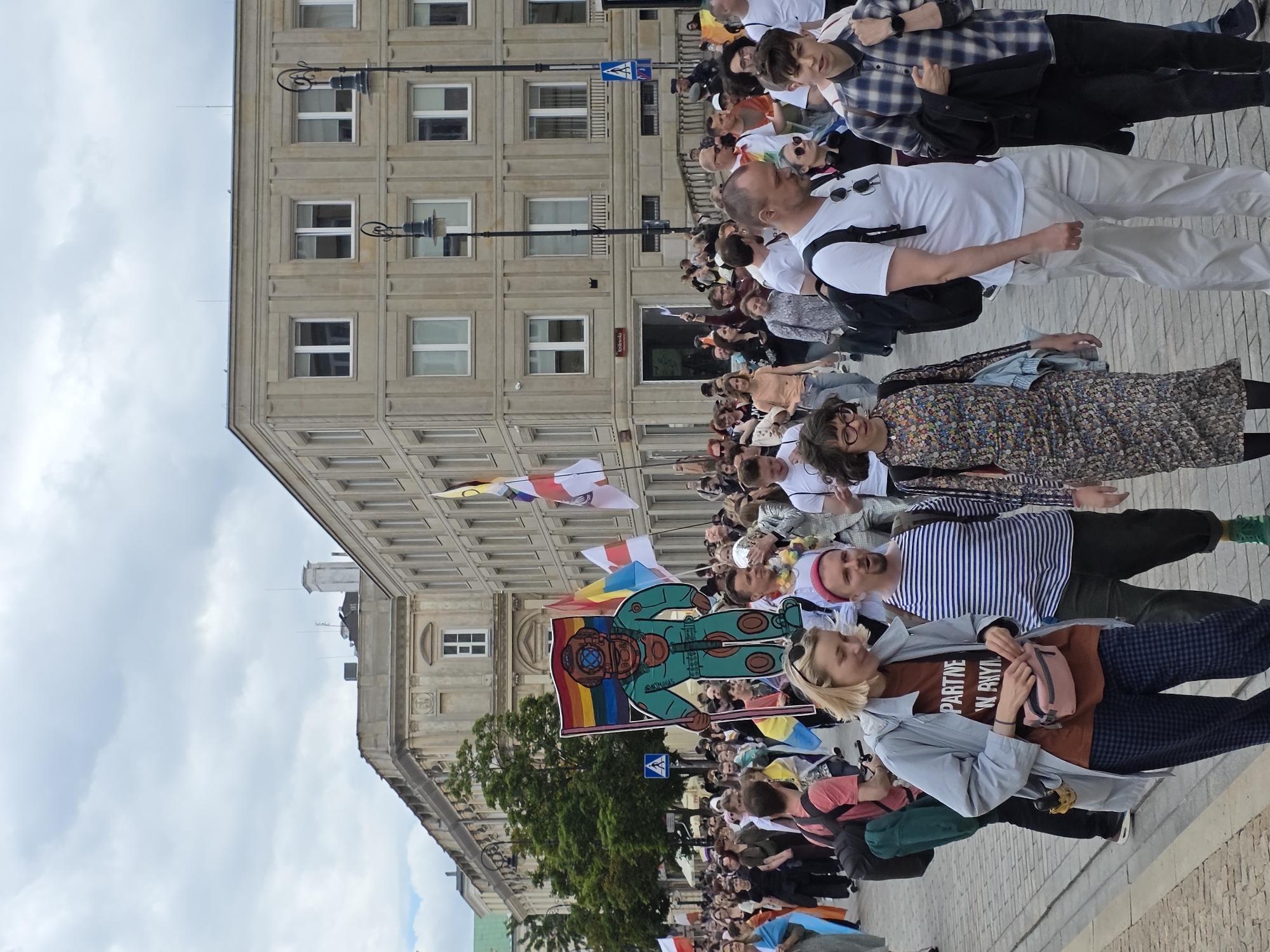 The image depicts a street scene in Warsaw, Poland, likely during a parade or public gathering, given the presence of many people and flags. In the foreground, a diverse group of individuals are walking, some looking at the camera, others engaged in conversation. A woman in a light blue jacket and checkered pants is prominently featured, as is a man in a white t-shirt and light-colored pants.
Behind them, a large crowd stretches into the distance, with many holding flags and banners. One notable banner features a cartoonish depiction of a diver in a green suit, holding a rainbow flag. Buildings with multiple windows line the street, suggesting an urban environment. The sky is overcast. The overall atmosphere appears to be one of community and shared activity.