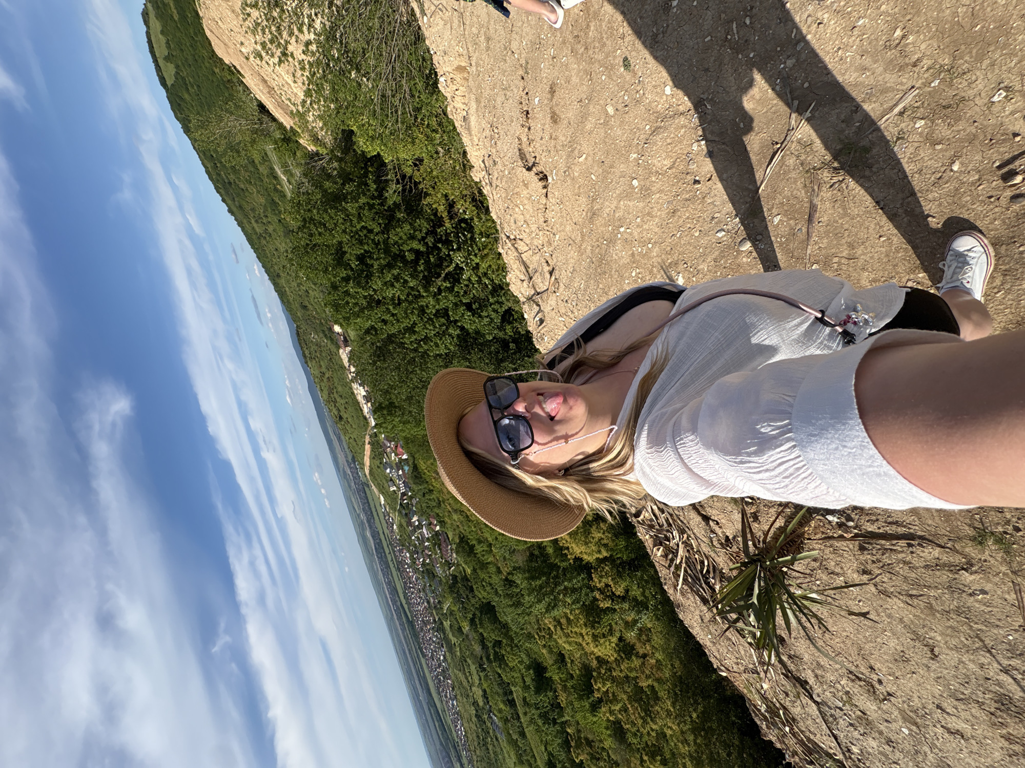 A selfie captures a woman on a high vantage point overlooking a vast landscape under a clear, bright sky. The woman, with blonde hair, wears a straw hat, sunglasses, and a white, crinkled long-sleeved top, with black shorts or a skirt visible. A delicate beaded chain is around her neck, attached to her sunglasses. She has a playful expression, sticking her tongue out slightly. Her shadow, along with the shadows of at least one other person, is cast prominently on the dry, stony ground, indicating bright midday or early afternoon sunlight.
The setting appears to be a cliff edge or a hilltop path made of earth and small rocks, with sparse, spiky green plants growing. To her left (viewer's left), the terrain drops off steeply, revealing a dense canopy of lush green trees that cover rolling hills. Beyond the trees, a wide valley stretches into the distance, dotted with numerous buildings suggesting a town or village. The horizon is visible, meeting a sky that transitions from a light blue near the horizon to a deeper blue overhead, adorned with elongated white clouds. In the upper right corner of the frame, parts of another person's legs and feet are visible, one wearing a white sneaker and the other a white flip-flop. No visible text is present in the image.