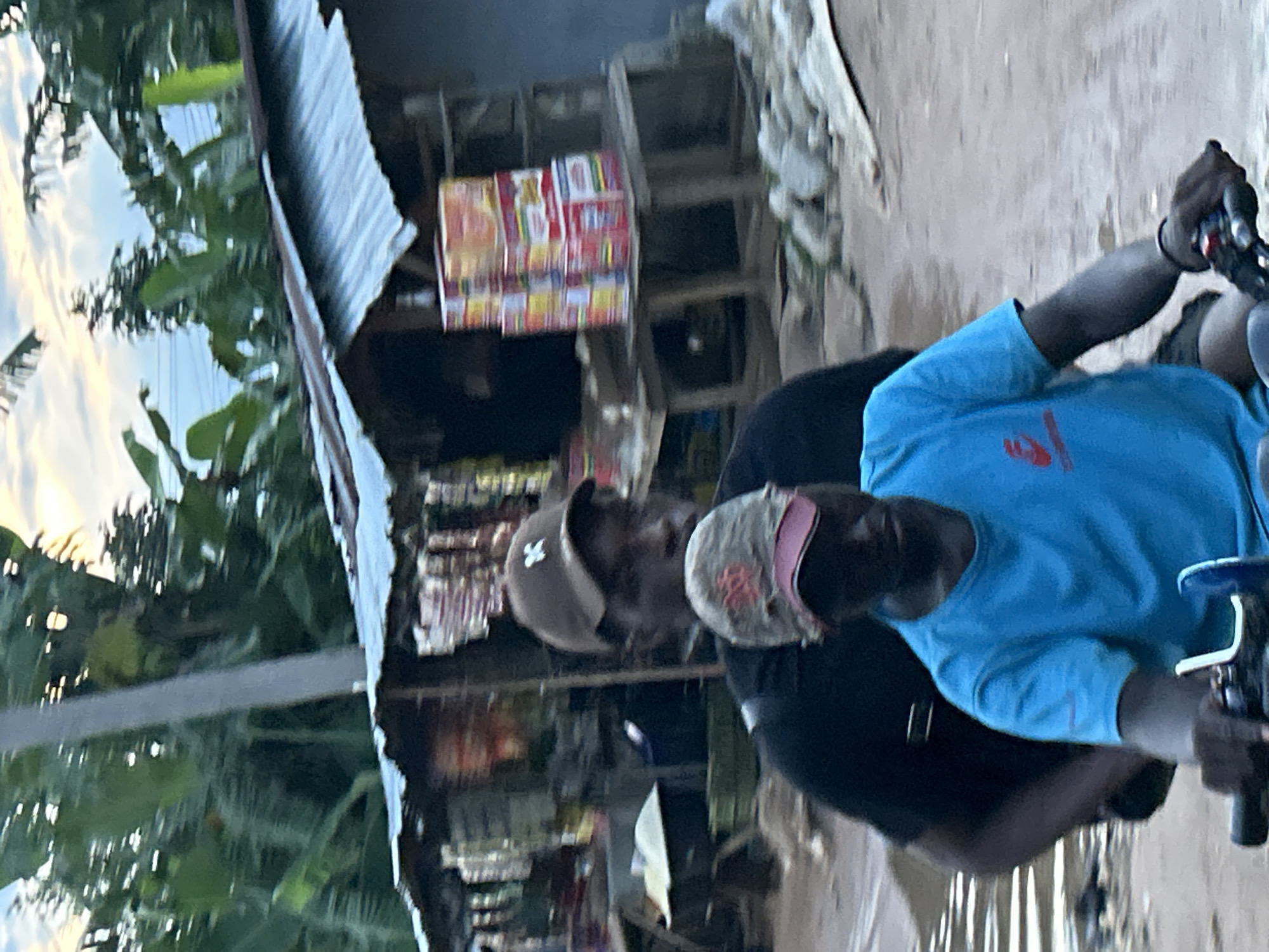 An outdoor scene captures two individuals on a motorcycle beside a roadside stall, likely in Ihiagwa, Nigeria. The image appears to be taken during daylight hours under a bright sky.
In the foreground, a person wearing a light-colored cap with a pink/red brim and a blue long-sleeved shirt is seated on a motorcycle. Their left hand is visible on the handlebars, which feature chrome elements. A red symbol or logo is present on the back of their blue shirt, but the text is not clearly discernible. Behind this person, a second individual is visible, wearing a brown or tan cap with a white symbol (possibly an 'M' or 'W') and a dark shirt, appearing as a passenger.
To the left of the individuals, a simple structure with a corrugated metal roof is visible, characteristic of a local shop or market stall. Inside this structure, various packaged goods, possibly snacks, drinks, or other sundry items, are stacked on shelves or displayed on a stand. These packages exhibit bright colors like red, white, and yellow, but no specific brand names or text are legible due to the distance and image resolution. Green foliage and trees are visible in the background, suggesting a natural environment surrounding the roadside. The ground appears paved or dusty.