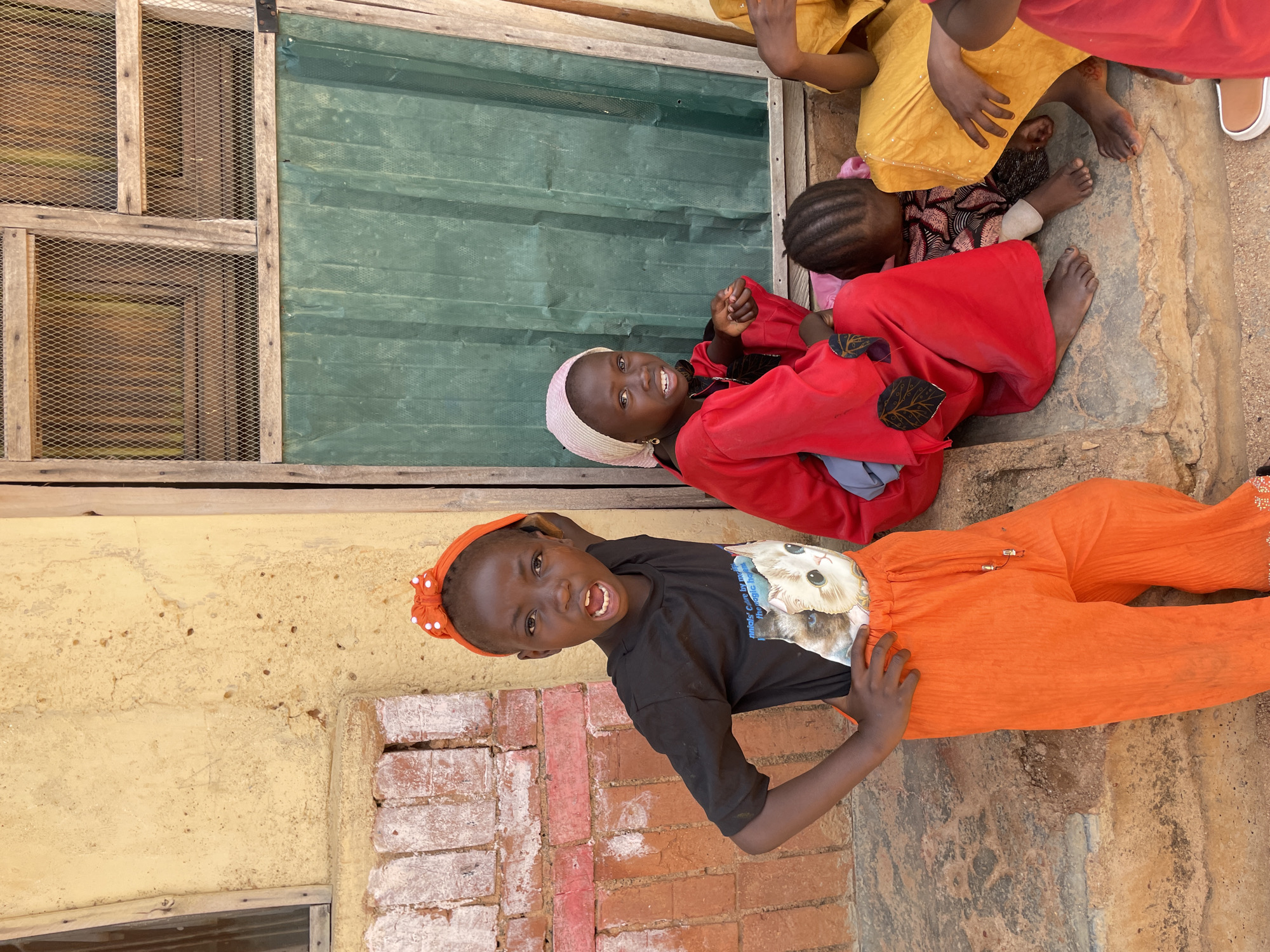 A medium shot captures four young children and a baby sitting on a concrete step outside a weathered building. The building features a yellow-painted wall with brick detailing and a wooden door with wire mesh windows and a green tarp. The child on the left, wearing an orange headscarf, black t-shirt with a cat print, and orange pants, strikes a dynamic pose with hands on hips, mouth open in an expression. Beside them, a child in a red garment and headscarf sits on the step, looking towards the camera. Another child is partially visible, leaning on the child in red, and a fourth child in yellow is seated to the right. The ground is a mix of dirt and concrete. The scene suggests a casual gathering in an urban environment, likely in Jos, Nigeria, given the architectural style and visible elements.