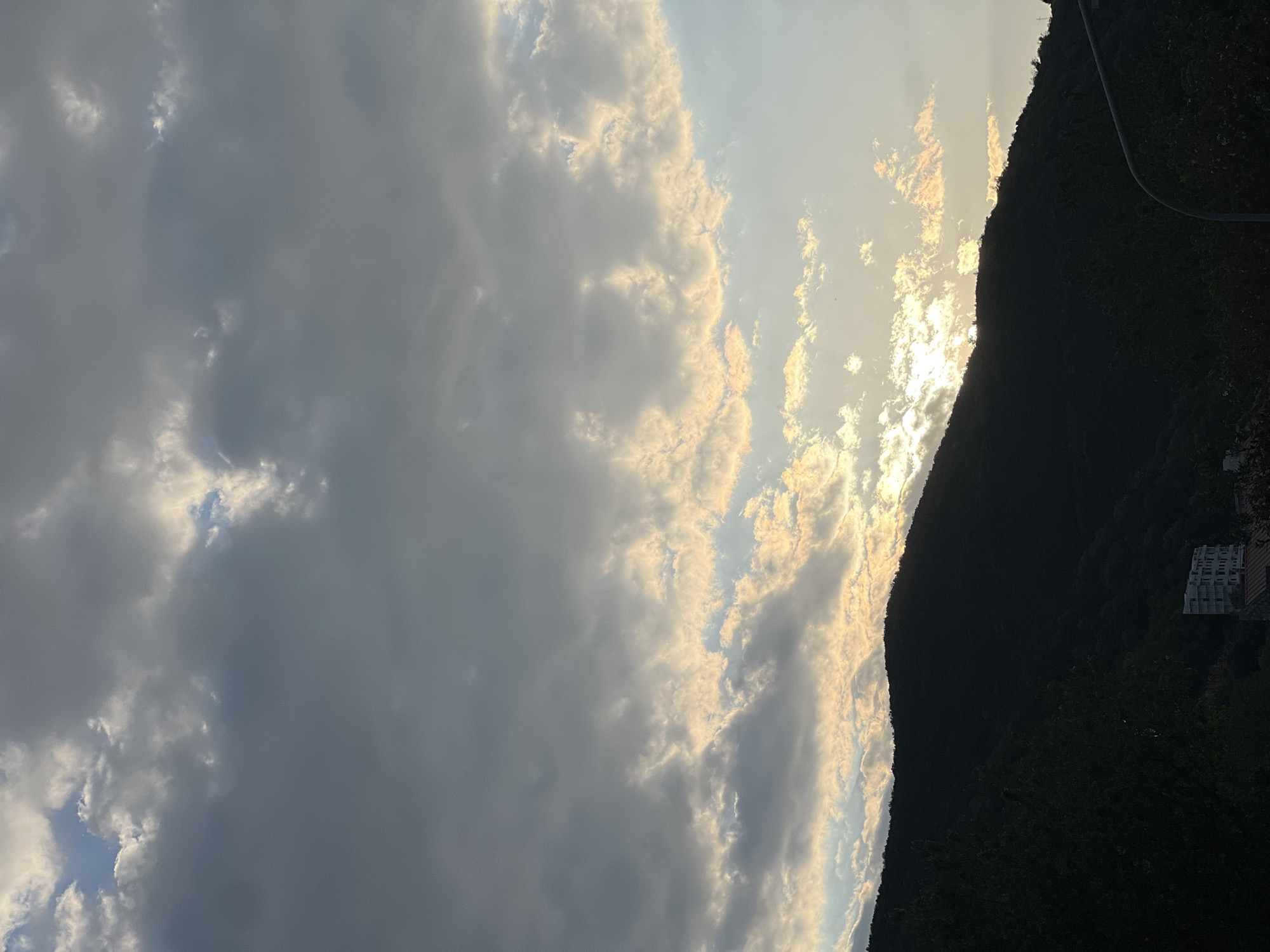 The image shows a landscape scene, possibly near the city of Budva, Montenegro. The sky is filled with a mix of dark and light clouds, hinting at either an approaching storm or a sunset. A sunlit area peeks through the clouds, casting a golden glow. The silhouette of a densely vegetated mountain range dominates the right side of the frame. On the mountainside, a building complex, possibly a hotel or residential area, is visible. A winding road snakes along the upper ridge of the mountain.