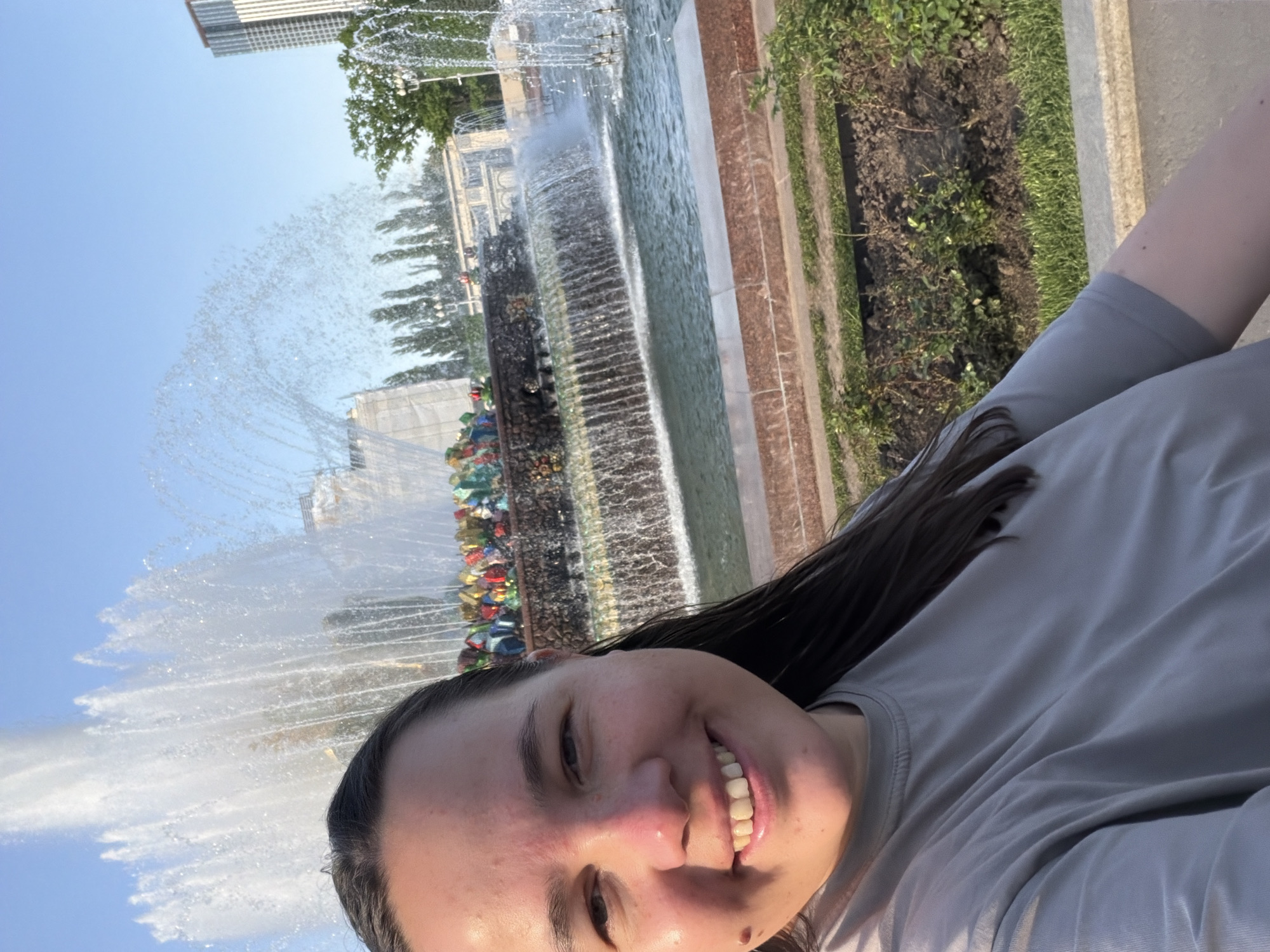 A selfie depicts a woman with dark hair wearing a light grey shirt, smiling with teeth showing. Behind her is a fountain with water spraying high into the air. The fountain is decorated with colorful elements. Behind the fountain, trees, buildings, and a blue sky are visible. The fountain is likely at VDNKh in Moscow, Russia.