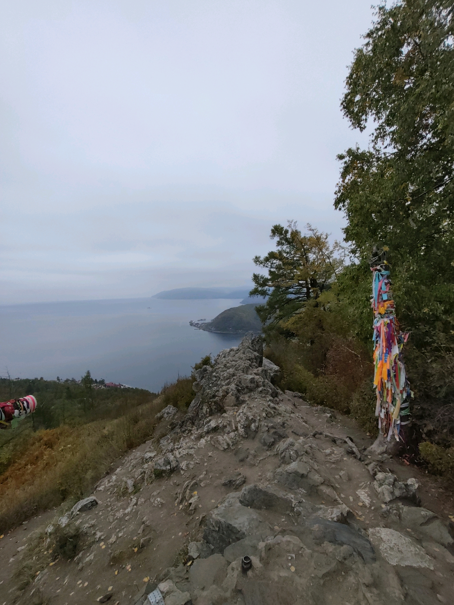 The image depicts a scenic overlook on a rocky terrain, likely in the vicinity of Nikola, Russia. A calm body of water, possibly a lake or sea, stretches towards the horizon under an overcast sky. The foreground is dominated by a rugged, rocky path leading towards the water. To the right, a tree trunk is adorned with numerous colorful ribbons, suggesting a cultural or spiritual significance. Vegetation, including trees and shrubs, is visible on the slopes leading down to the water, with some areas displaying autumn colors. In the distance, a shoreline with structures and hills can be observed. A fragment of what appears to be a piece of clothing with a colorful pattern is visible in the lower left corner of the image. The overall atmosphere is serene and natural, with a hint of human presence indicated by the ribbons and the distant settlement.
