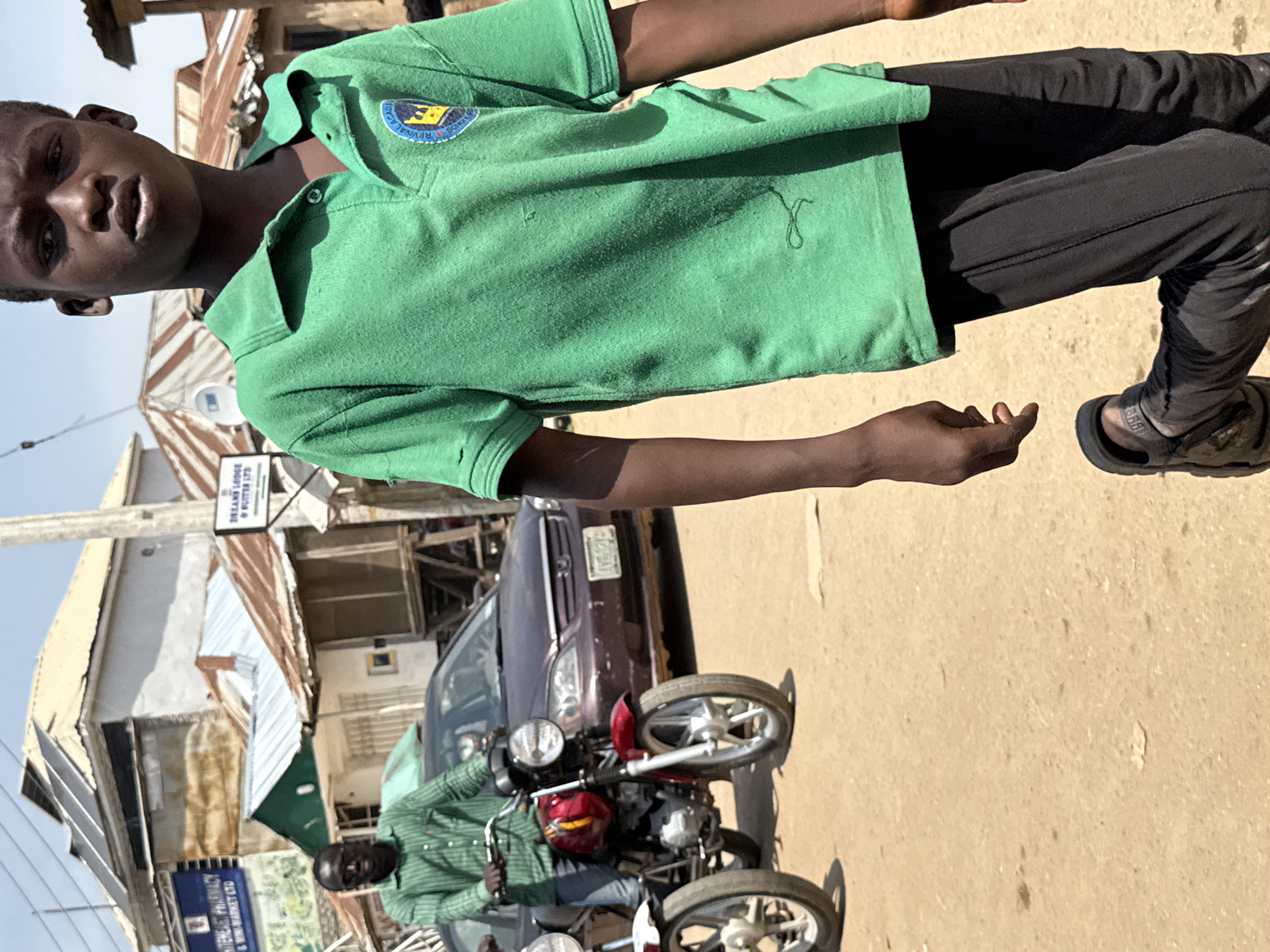 In an outdoor urban setting in Abuja, Nigeria, a young person in a green polo shirt and black pants is in the foreground, facing right and slightly away from the camera, with their left leg bent as if in motion. Behind them, a man in a green striped shirt rides a red motorcycle, with another motorcycle partially visible to the left. A dark sedan is parked behind the man on the motorcycle. Buildings with corrugated metal roofs and some with solar panels are visible in the background, suggesting a mix of residential and commercial structures typical of some areas in Abuja. The ground is a dusty, unpaved surface. The overall atmosphere is casual and appears to be a street scene with moderate activity.