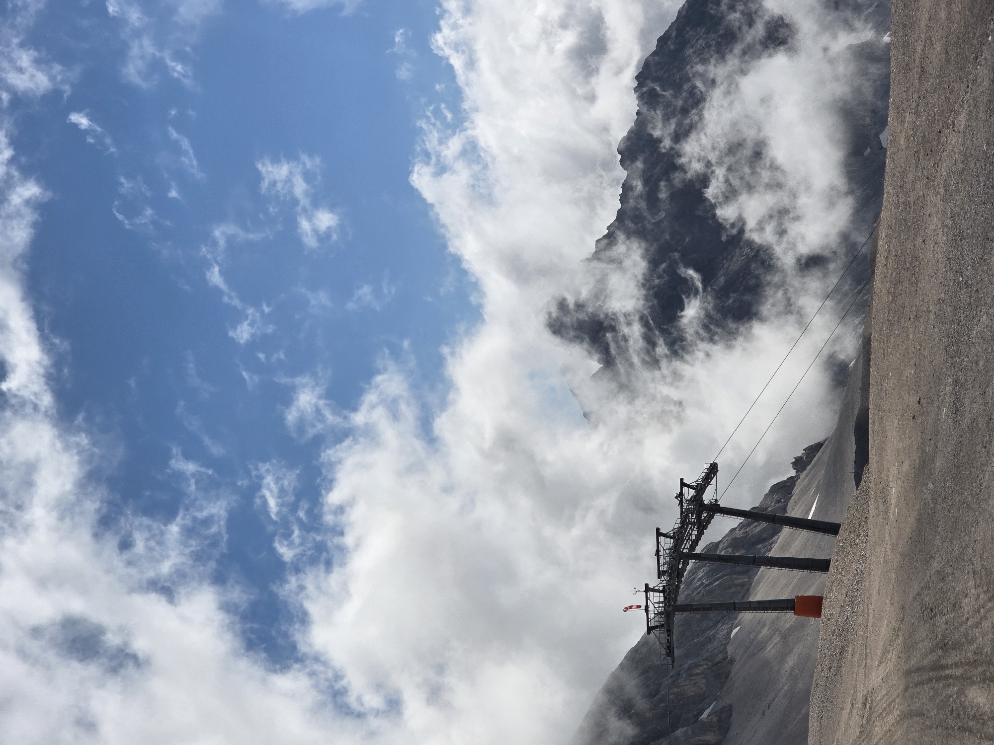 The image depicts a mountain landscape with a ski lift in the foreground. The ski lift consists of three large metal towers supporting cables. One of the towers has an orange base. At the top of the towers, there is a red and white windsock. The ground in front of the ski lift is a gravelly, dusty surface with tire tracks. Behind the ski lift, steep, rocky mountains rise, partially obscured by white clouds. The sky is blue with scattered white clouds. This scene is characteristic of the Zugspitze, Germany's highest peak, located near Garmisch-Partenkirchen. There are no people visible in the image.