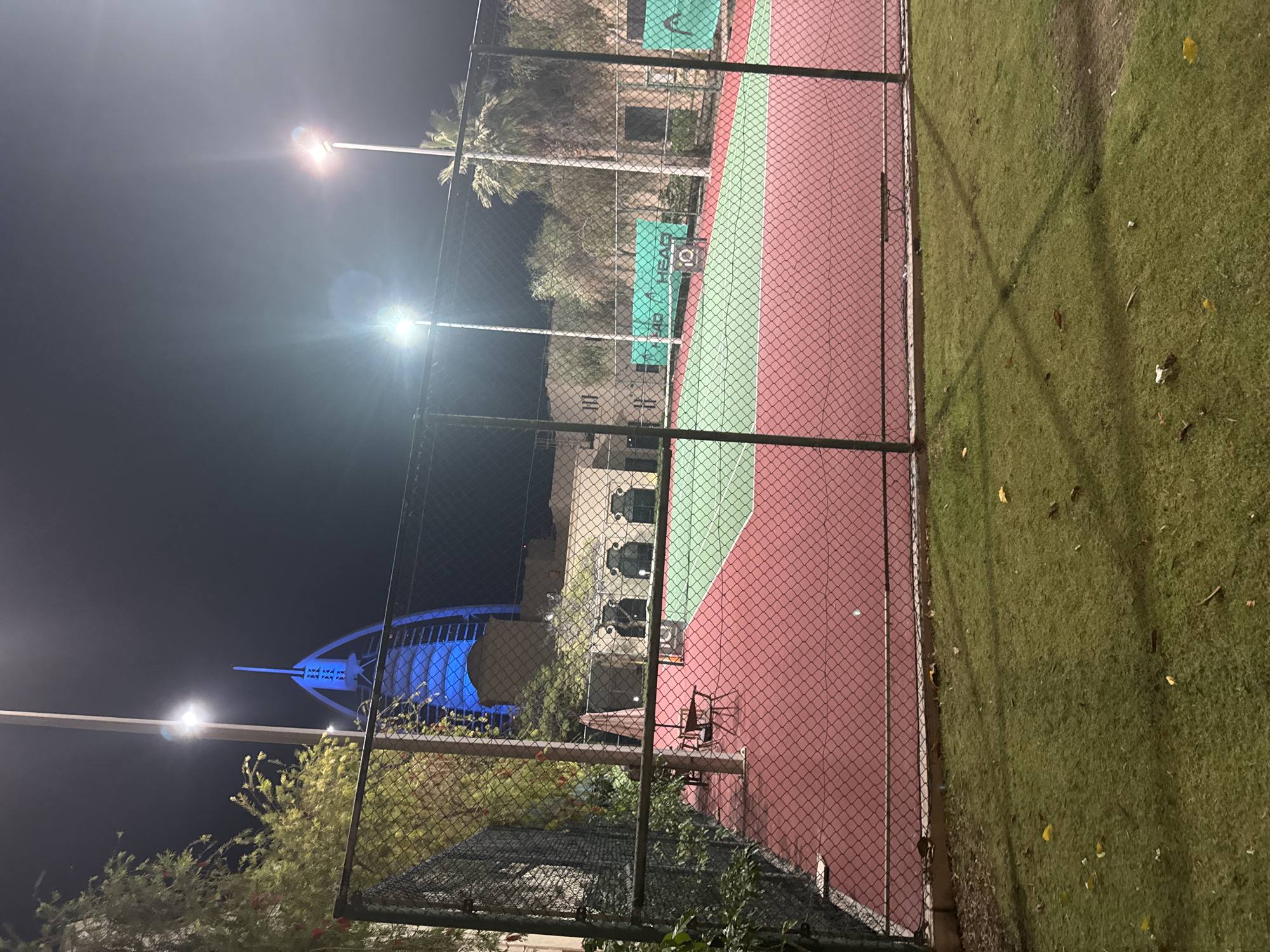 Here is a description of the image:
The image shows a tennis court in Dubai, United Arab Emirates, at night. The Burj Al Arab hotel, illuminated in blue, is visible in the background. The tennis court is enclosed by a chain-link fence. The court surface is red and green. There are light poles around the tennis court. Grass surrounds the outside of the fence line on the right side of the frame.