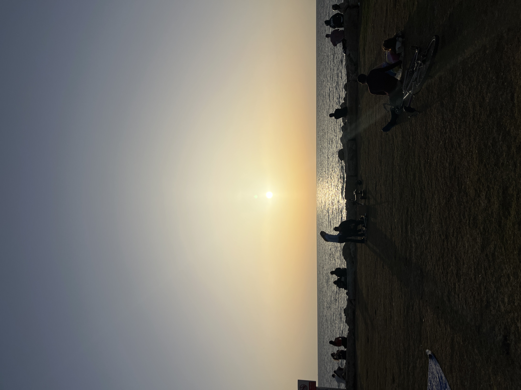 The image shows a sunset scene along the Tel Aviv coastline in Israel. The sun is setting over the Mediterranean Sea, casting a bright glow on the water and creating silhouettes of people gathered along the shore. There are individuals sitting and standing along the rocks, as well as a bicycle parked on the grassy area adjacent to the coast. In the lower part of the frame, what appear to be a No Smoking sign and the edge of a sunshade are visible. The sky transitions from a bright yellow-orange near the sun to a cooler blue-gray further away.
