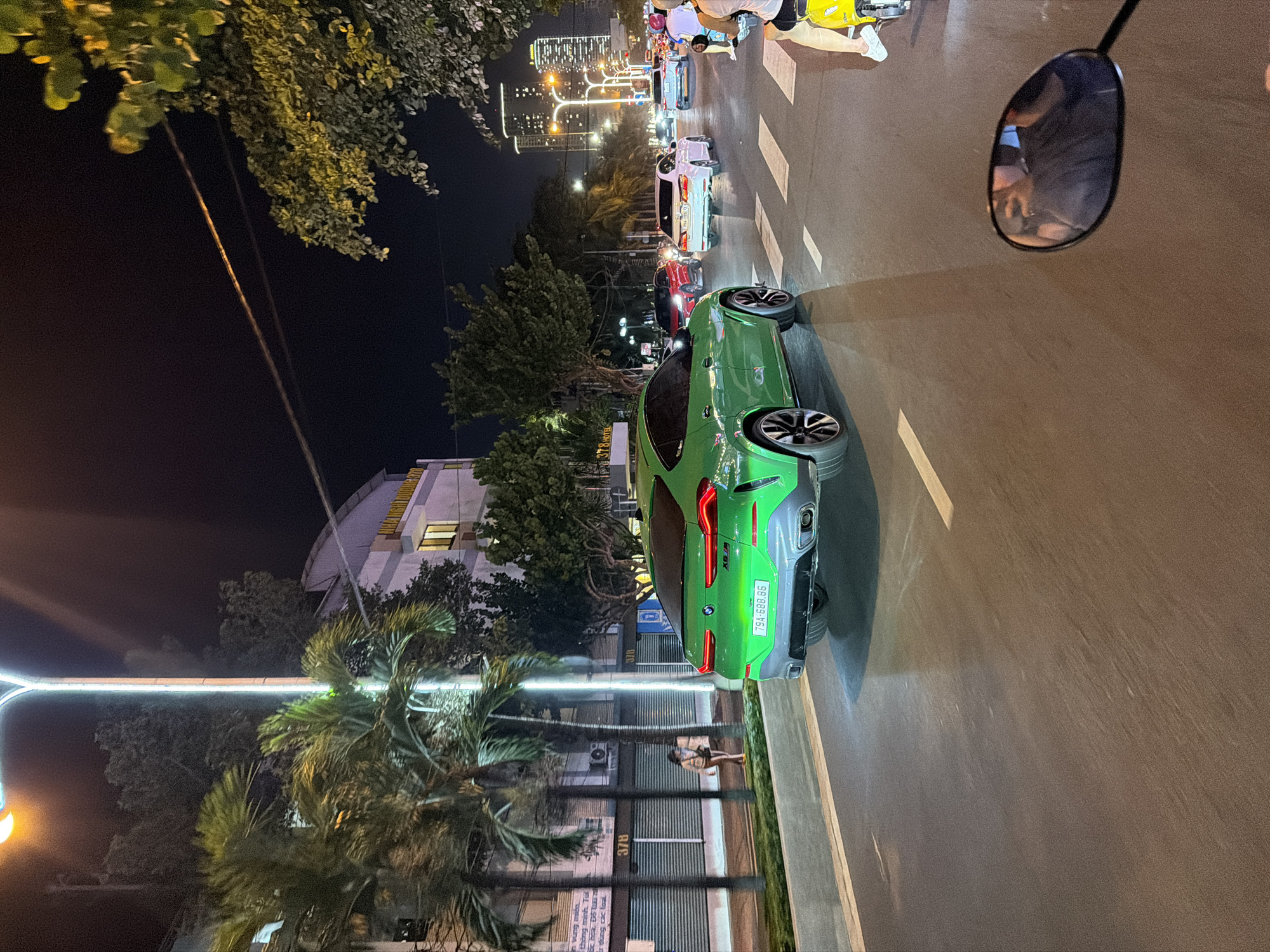 The image depicts a nighttime street scene in Nha Trang Ward, Vietnam. In the mid-ground, a bright green BMW X6 M SUV, bearing Vietnamese license plate "79A-688.86", drives away from the viewer on an asphalt road. Its rear lights are illuminated, and the vehicle features a grey lower diffuser with visible quad exhaust tips.
In the immediate foreground, to the bottom right, the scene is viewed from a yellow scooter, indicated by the partial visibility of its rear-view mirror reflecting a person holding a smartphone, and the lower legs of the rider wearing white sneakers. The asphalt road surface exhibits white dashed lane markings and a pedestrian crosswalk, with sections appearing reflective, possibly due to moisture or artificial lighting.
Behind the green BMW, a white sedan with license plate "79A-196.17" follows, and a red SUV is visible further down the road on the left. Multiple other vehicles, including cars and motorbikes, are present in the distance, suggesting moderate urban traffic.
On the left side of the street, a paved sidewalk is bordered by several palm trees and a prominent vertical streetlight emitting white LED illumination. Behind these, a multi-story building is visible, displaying "378" and "378 HOTEL" signage. A pedestrian, dressed in a light-colored jacket and dark pants, walks away from the camera on this sidewalk.
The background reveals an urban environment with various buildings lining the street. In the far distance, a cluster of illuminated high-rise buildings forms the city skyline, with "ASIA HOTEL" faintly visible on one structure. The overall scene is lit by numerous streetlights, casting bright light onto the vehicles, foliage, and building facades. Trees and dense foliage are present along both sides of the street. The sky is dark, consistent with nighttime conditions.