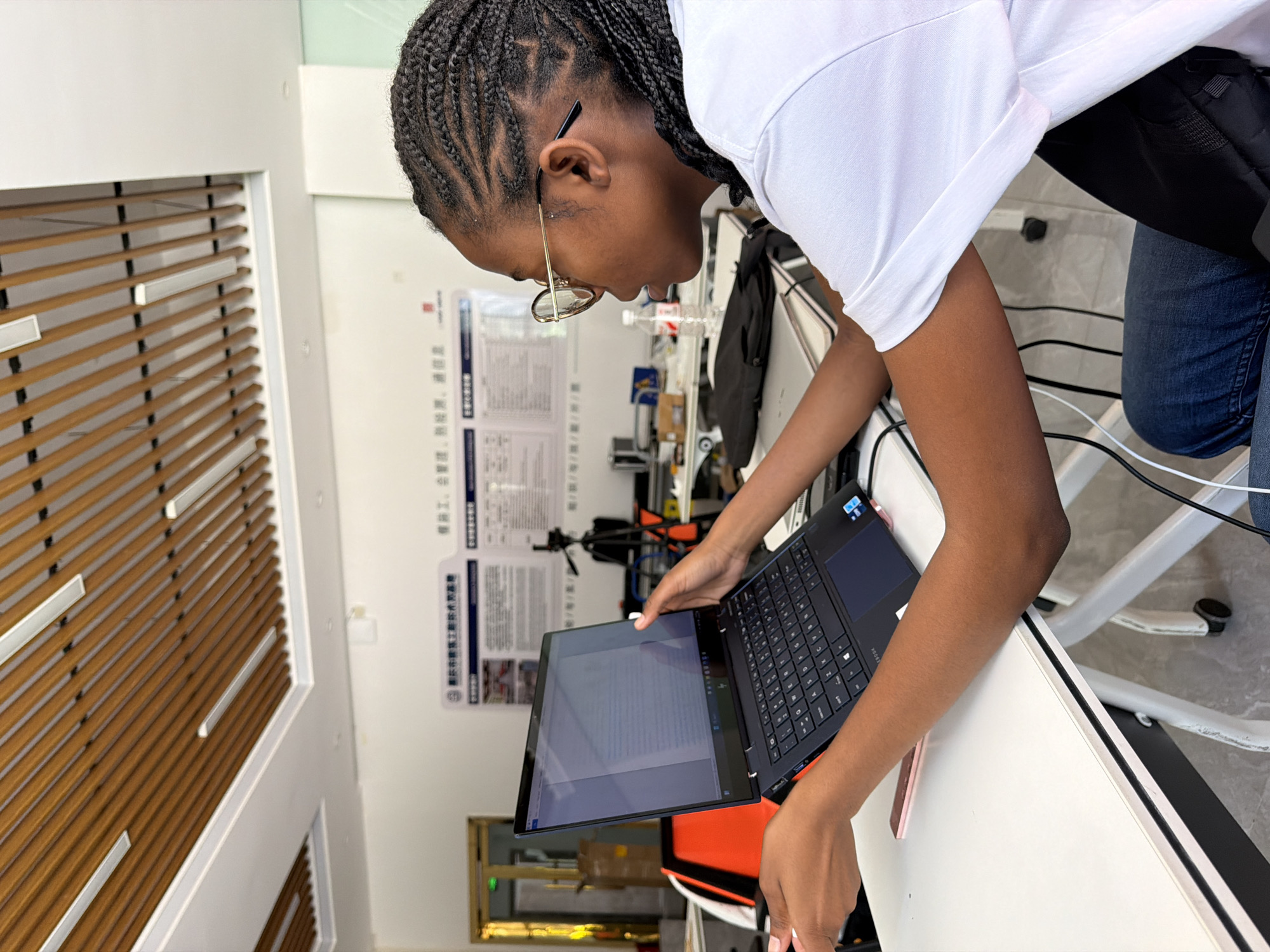 In Jiangjin, China, a young Black woman with braided hair and glasses is seated at a white desk, intently working on a dark blue laptop. The laptop is open and positioned on a stand, with its screen displaying text. Her hands are visible, one resting on the keyboard and the other near the screen, suggesting she is actively engaged in typing or navigating the content. She wears a white t-shirt and blue jeans. The background reveals a room with a white wall adorned with posters containing Chinese text and diagrams. A tripod is set up behind the desk, and a bottle of water is on the desk. The ceiling features a modern design with wooden slats and fluorescent lighting. The overall environment appears to be an office or workspace.