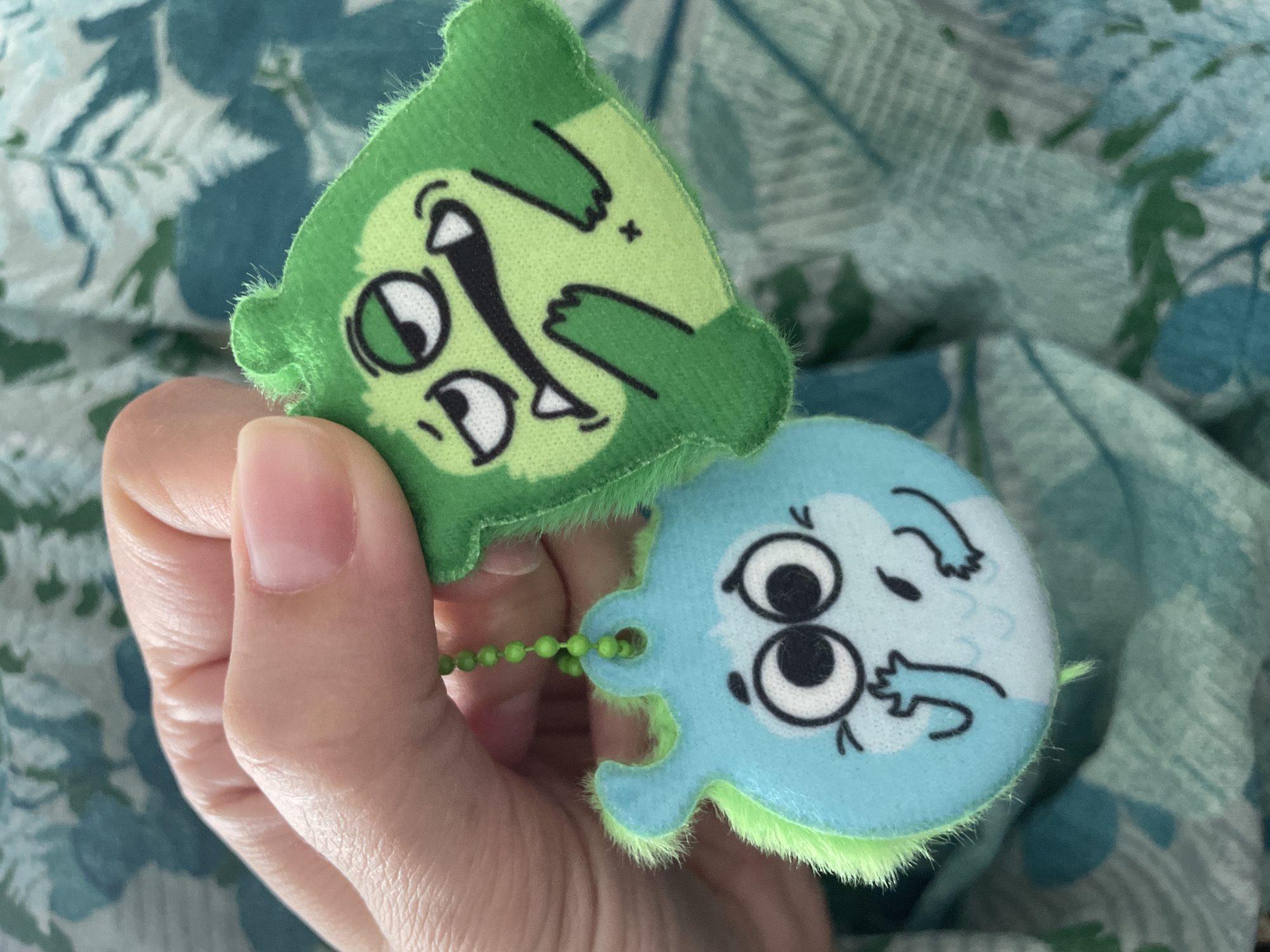 A hand is holding two plush keychains, one green and one blue, against a background of a blue and green patterned blanket. The green keychain has a cartoon face with exaggerated features, and the blue keychain has a similar style of face. The keychains are connected by a short green beaded chain. The setting is not Izhevsk, Russia as there is no information to indicate this.