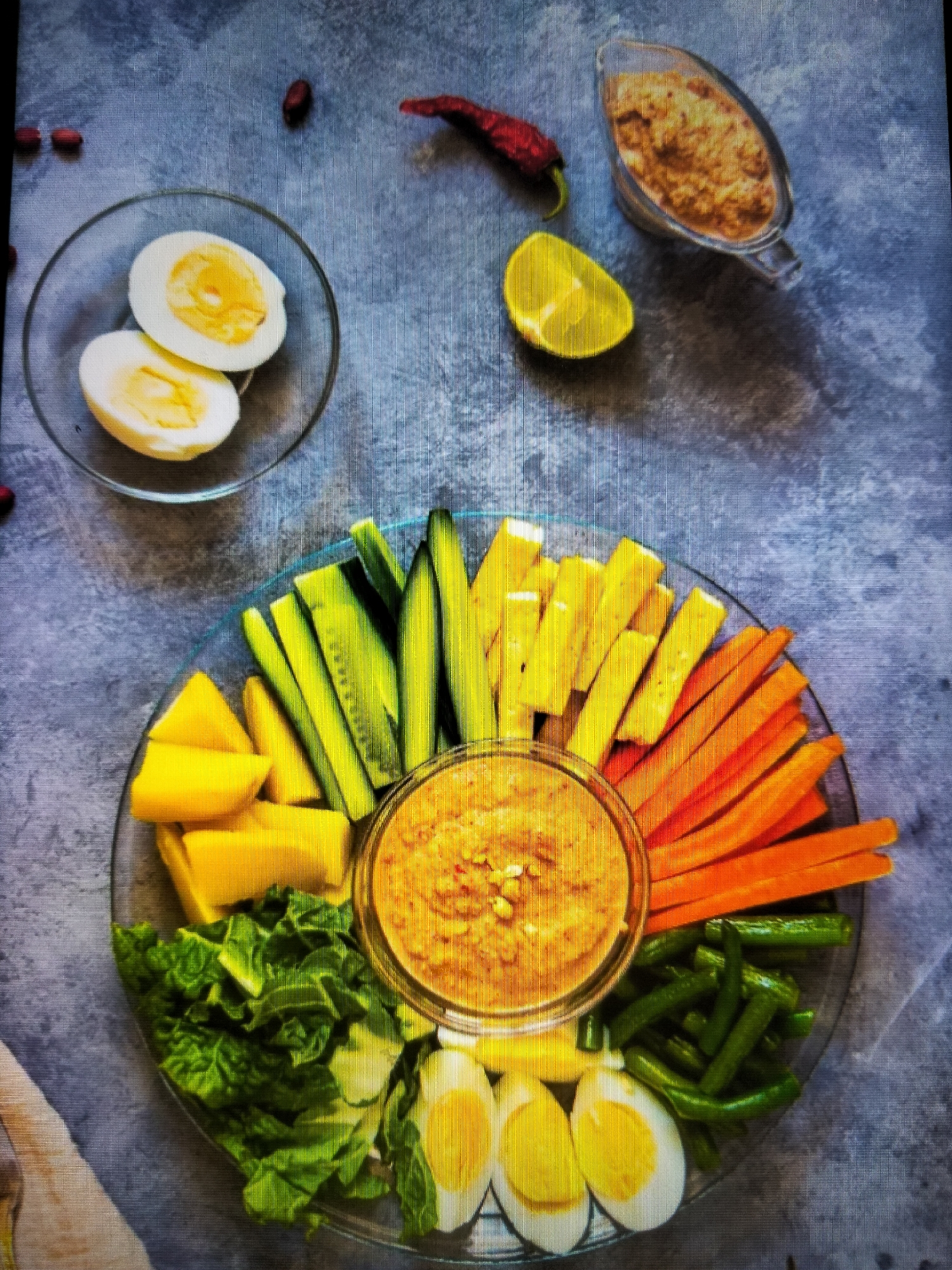 A platter of assorted fresh vegetables and hard-boiled eggs is presented with a side of peanut sauce. The platter is arranged in a radial pattern, with segments of cucumber, carrots, yellow bell peppers, green beans, lettuce, mango, and slices of hard-boiled eggs. In the center of the platter is a bowl of thick, textured peanut sauce. To the top right of the platter, a small gravy boat holds more of the peanut sauce, alongside a wedge of lemon and a dried red chili pepper. Two hard-boiled eggs, halved, sit in a small glass bowl to the top left. Several peanuts are scattered on the grey, textured surface beneath the platter. The scene is lit from above, casting shadows on the surface. The image is captured from a top-down perspective. There is no indication of the city of Payagaboh, Indonesia in the image.