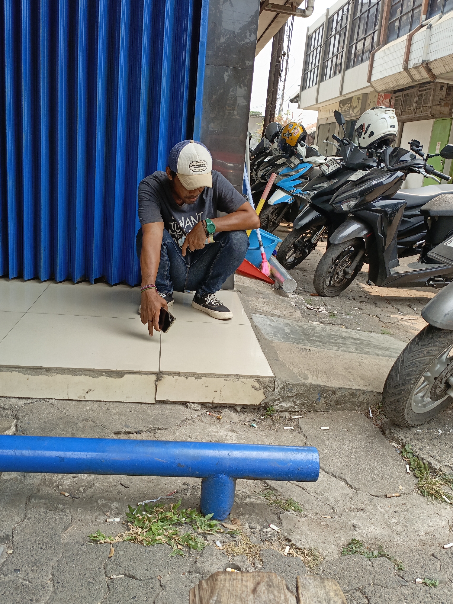A man is crouching on a tiled porch next to a blue corrugated metal roller door. He is wearing a grey t-shirt, blue jeans, a beige and white baseball cap, and sneakers. In his right hand, he holds a black smartphone, which he appears to be looking at. To the right of the man, several motorcycles are parked on a cracked concrete ground. One motorcycle has a white helmet on its handlebars. In the background, there is a multi-story building with windows. In the foreground, a blue pipe runs horizontally across the frame. The scene appears to be in an urban area, likely a street or alleyway, in East Jakarta, Indonesia, indicated by the common sight of parked motorcycles and the style of the building.