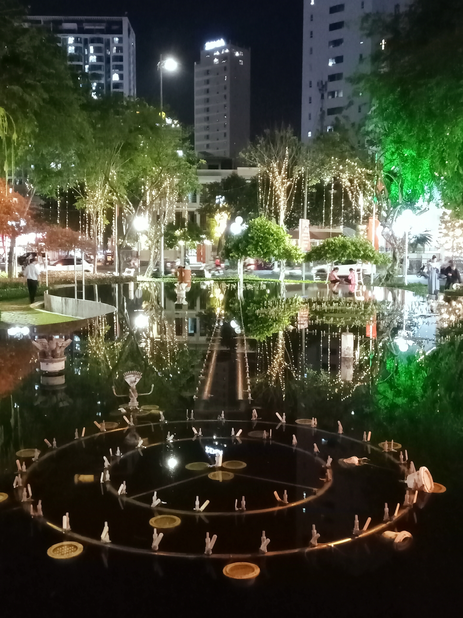 The image presents a vibrant night scene in an urban park, likely in Nha Trang Ward, Vietnam. A large, still body of water, possibly a decorative pond or fountain, occupies the foreground and midground, creating a striking mirrored effect. In the water, a circular or spiral arrangement of fountain nozzles or submerged lights is visible, with multiple individual elements projecting upwards from golden bases.
Along the perimeter of the water feature, numerous trees are brightly illuminated with strings of warm white lights, their reflections dancing on the dark surface. Some trees are also bathed in green light. In the midground, several people are visible, including a man in a white shirt standing near the water's edge on the left, and a few individuals sitting or standing on the far side of the water, seemingly relaxing and enjoying the evening atmosphere. Their reflections are also clearly visible in the water.
In the background, multiple tall, modern buildings rise against the dark night sky, their windows glowing with internal lights. One building to the far right features a prominent sign with white letters at its peak, which appears to spell "SOVIMA". Streetlights further illuminate the scene, adding to the urban nighttime ambiance. The overall impression is one of a peaceful yet lively public space at night, enhanced by decorative lighting and the reflective quality of the water.