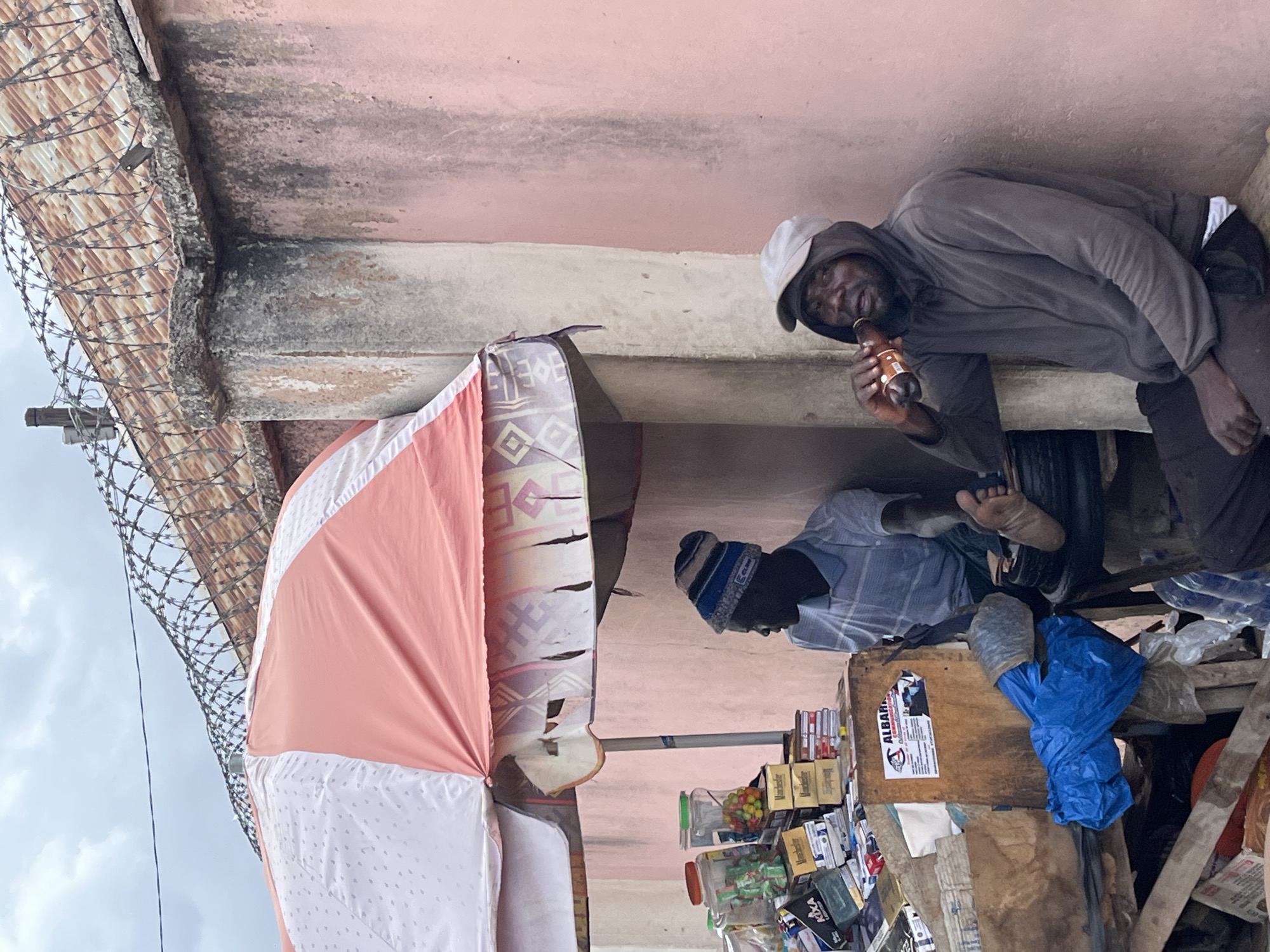 This image depicts an informal street scene, likely a market stall, in Jos, Nigeria, during daytime under an overcast sky. Two men are featured.
On the right, a dark-skinned man in a grey hooded sweatshirt and a light-colored cap is seen drinking from a brown bottle, looking towards the viewer with a slight smile. He has a beard and appears relaxed.
Further to the left, another dark-skinned man, wearing a striped blue and white beanie and a light blue striped shirt, is seated or crouched behind a wooden stall. His bare foot is visible near the stall's base. The stall functions as a small shop, displaying various items for sale, including small plastic containers of colorful sweets or candies and multiple boxes of "Manchester" cigarettes. Other visible packaging includes "MXA" and "KICA" branded items. A blue plastic bag rests on the stall, which also features a sticker partially visible, likely reading "ALBAINA".
Above the scene, a pink-painted wall and a concrete beam form the background. To the far left, corrugated iron roofing topped with coiled barbed wire fencing indicates a boundary or security measure. A large pink and white striped fabric, possibly an awning or umbrella, provides shade over part of the stall. The overall setting suggests a bustling, somewhat rustic urban environment.