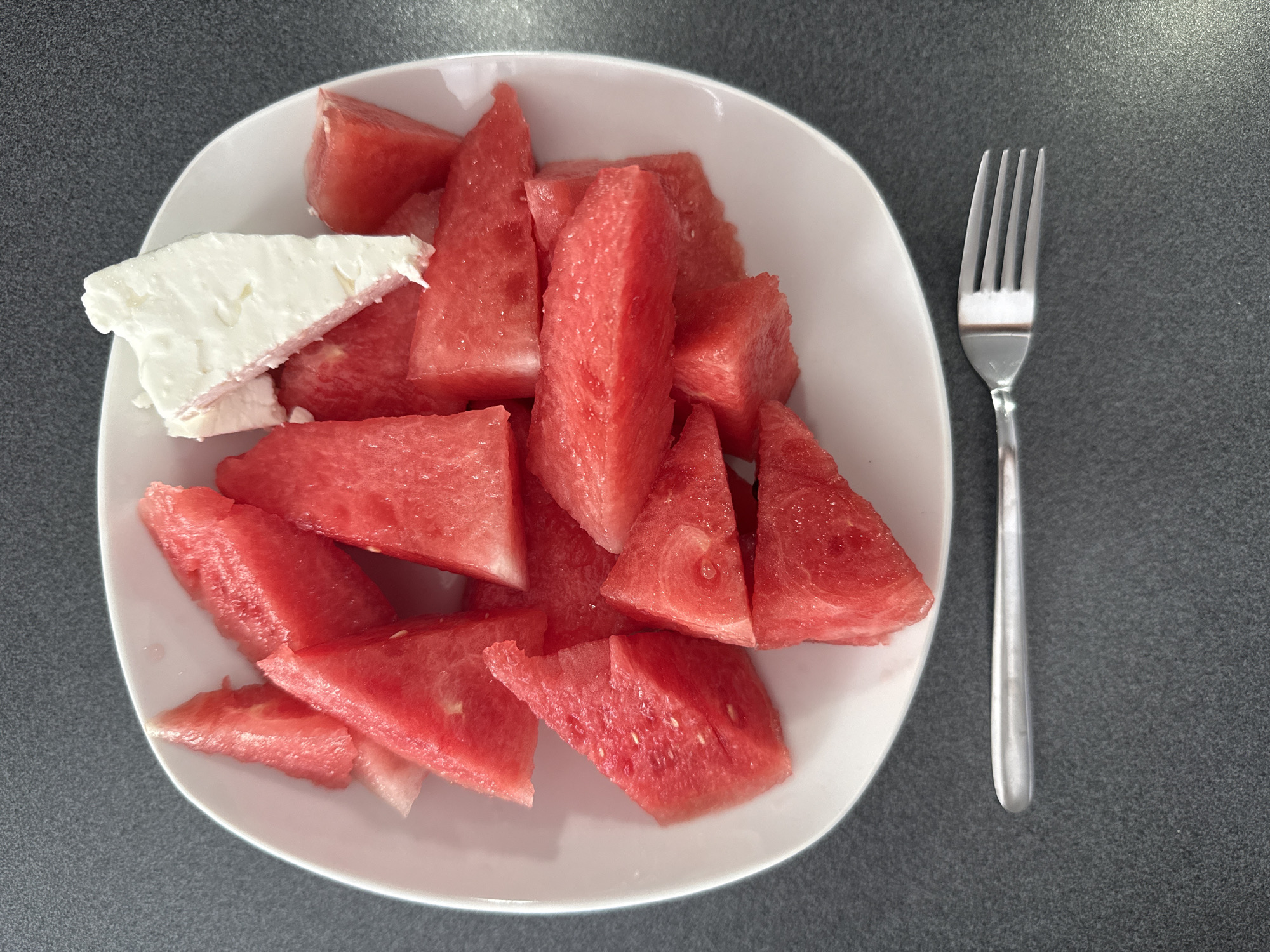 A white plate filled with sliced watermelon and feta cheese sits on a dark gray speckled surface. A silver fork rests below the plate. The watermelon slices are triangular and vary in size, with some pieces showing seeds. The feta cheese is cut into a wedge shape and is white and crumbly. The scene appears to be an indoor setting, possibly a kitchen or dining area, given the table surface and the presence of a fork. There are no people or animals visible in the image, and no specific landmarks or details that would indicate the location in Marum, Netherlands.