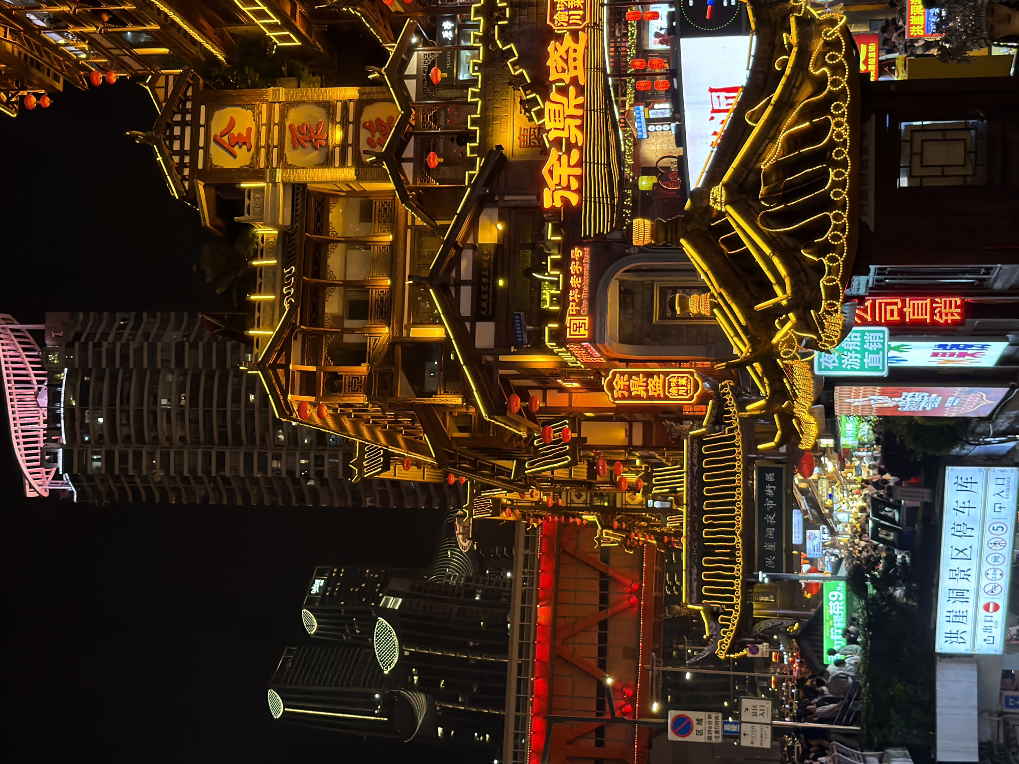 The image captures a vibrant nighttime scene in the Yuzhong District of Chongqing, China, likely at the Hongyadong tourism area. The architecture is a blend of traditional Chinese multi-story buildings, adorned with intricate wooden details and illuminated by warm yellow and red lights. These traditional structures are juxtaposed against a backdrop of modern skyscrapers, some of which are also lit up, creating a striking contrast between old and new.
The foreground features a detailed view of the traditional architecture, with its characteristic tiled roofs and ornate decorative elements. Numerous red lanterns hang from the buildings, adding to the festive atmosphere. Brightly lit signs with Chinese characters are visible, advertising businesses and attractions. One prominent sign in the mid-ground reads "渝景盛" (Yujing Sheng).
Below the traditional buildings, a bustling street scene unfolds, filled with a large number of people. They appear to be walking, shopping, and enjoying the nightlife. The street is also lined with shops and stalls, further enhancing the lively ambiance. In the lower part of the image, a sign for "洪崖洞景区停车场" (Hongyadong Scenic Area Parking Lot) is clearly visible, indicating the location's function as a tourist hub.
The overall environment is dark, as expected for nighttime, but the abundant artificial lighting from the buildings, signs, and lanterns creates a dynamic and visually rich composition. The image conveys a sense of energy and activity, characteristic of a popular urban tourist destination at night.