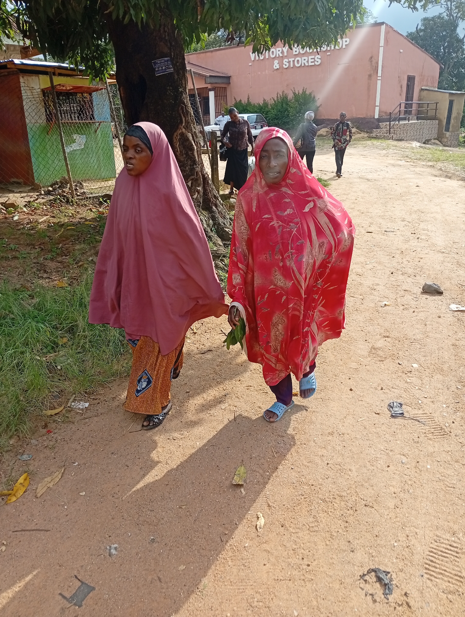 Two women are walking on a dirt path in front of a building with "VICTORY BOOKSHOP & STORES" written on it. The woman on the left is wearing a dark headscarf and a rose-colored abaya over a patterned skirt. She is looking towards the camera. The woman on the right is wearing a red patterned abaya and blue sandals. She is looking forward. Behind them, there are more people walking and a white vehicle. The environment is rural with trees and grass visible.