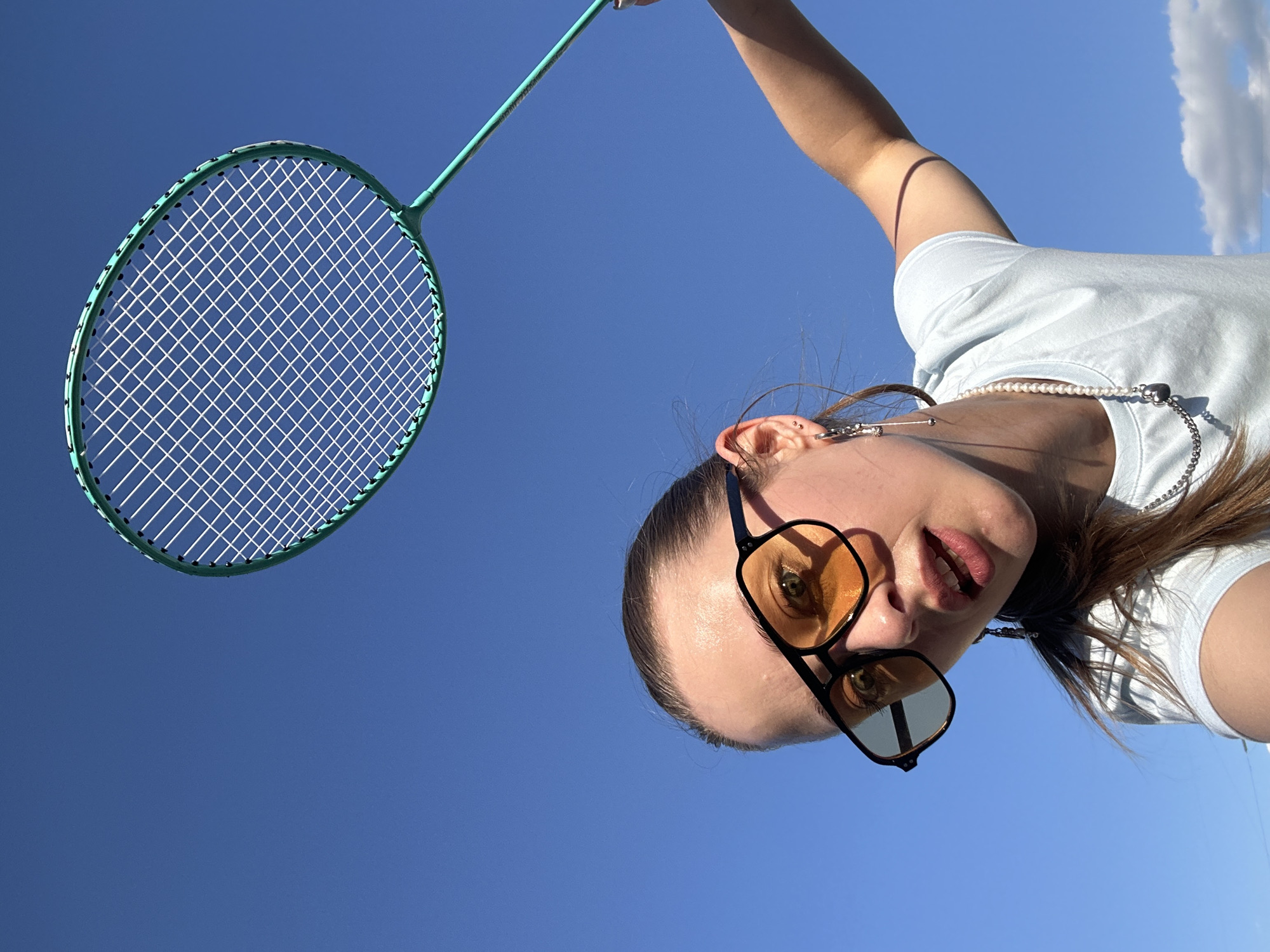 In a low-angle shot against a clear blue sky, a young woman is positioned in the foreground, her face turned towards the camera. She is wearing sunglasses with amber-tinted lenses and a light blue t-shirt. A delicate pearl necklace is visible around her neck. Her right arm is extended upwards, holding a badminton racket by its handle. The racket's strings and green frame are sharply in focus, set against the vast expanse of the sky. A single fluffy white cloud is visible in the upper right corner of the frame. The lighting suggests it is a bright, sunny day, possibly late afternoon due to the angle of the light casting a shadow on the woman's arm. The setting appears to be outdoors, likely in a park or recreational area in Khimki, Russia, given the context. The woman's expression, with slightly parted lips and wide eyes, conveys a playful or perhaps enthusiastic mood. There is no visible text in the image.