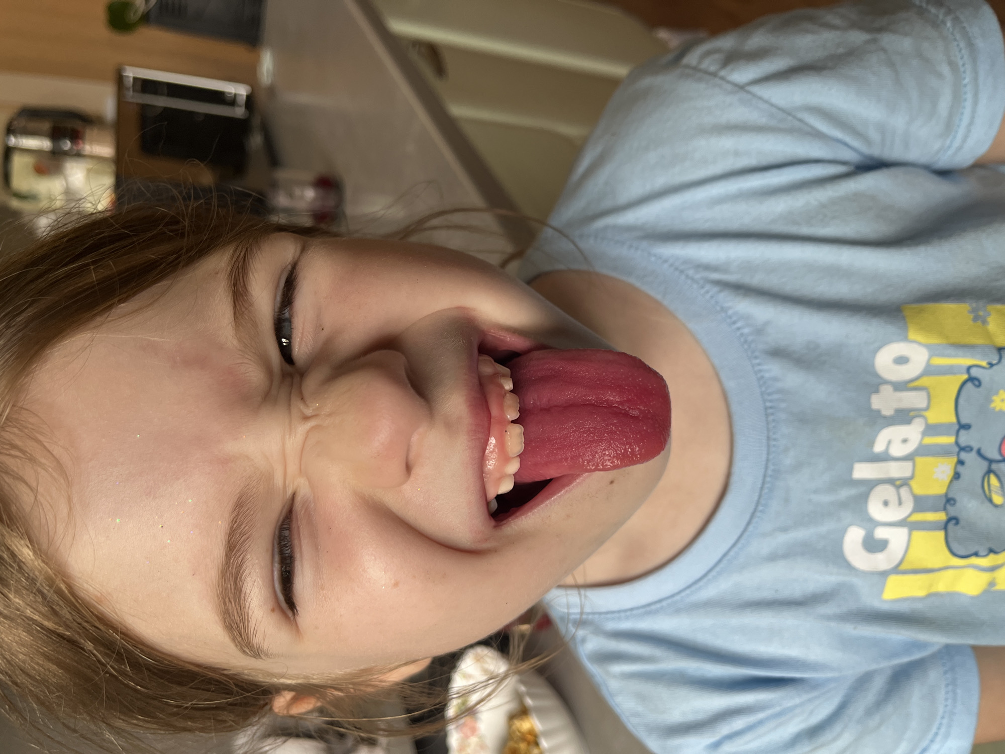 A close-up, eye-level, medium shot captures a young girl with light brown hair sticking out her bright red tongue. She is squinting her eyes and wrinkling her nose, making a funny face. She wears a light blue t-shirt with the word "Gelato" printed on it in yellow and white letters. The background is blurred but appears to be a kitchen with cabinets and appliances. The girl's face is lightly dusted with glitter on her forehead and cheeks. The image was taken indoors, likely in a home kitchen, with artificial lighting. There is no discernible information in the image that would place it in Sochi, Russia.