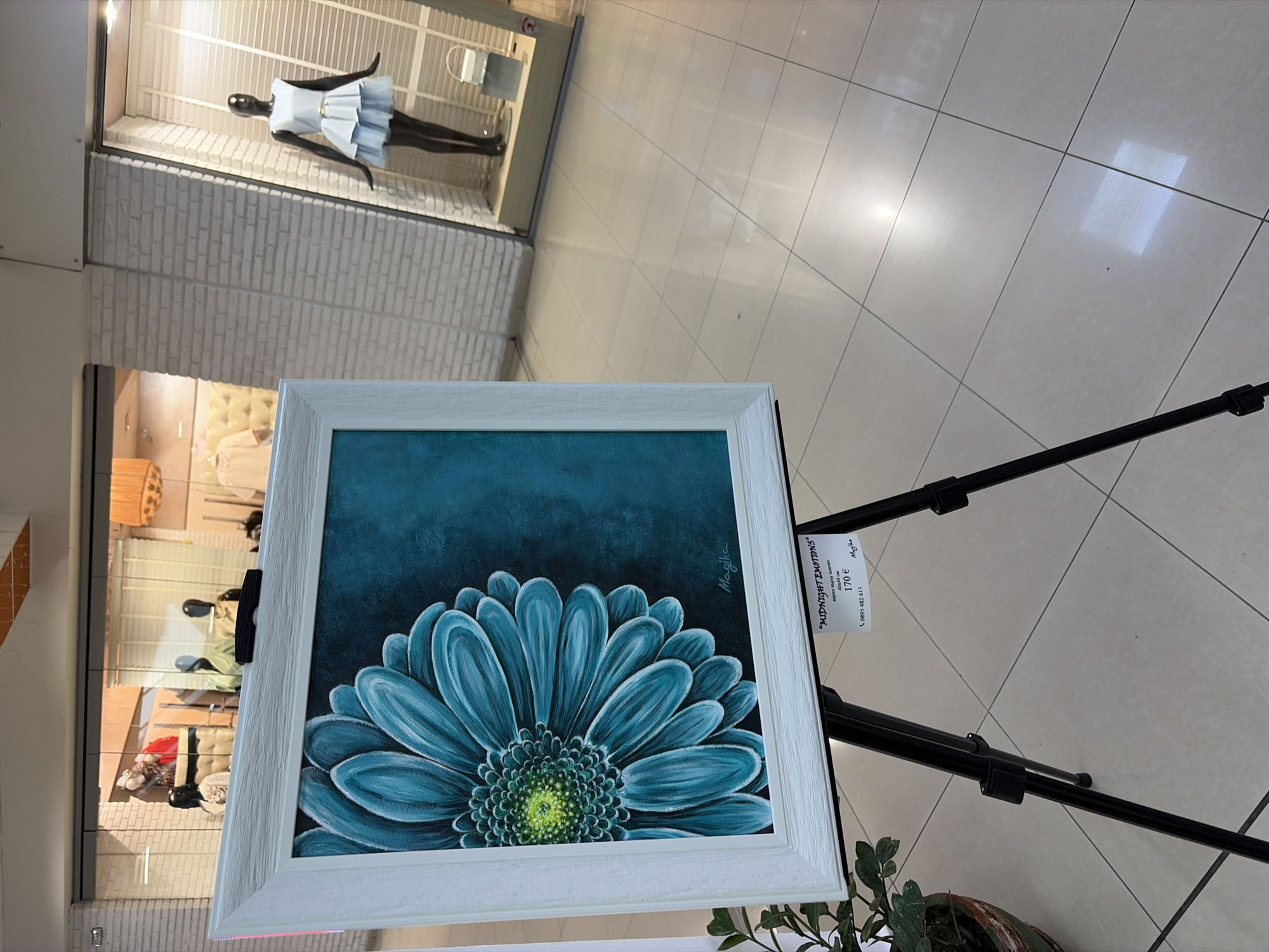 The image captures a scene within a shopping mall or commercial space, likely in Plovdiv, Bulgaria, showcasing an art exhibit with a flower painting prominently displayed on an easel. The painting, featuring a blue flower with a dark blue background and a white frame, is titled "MIDNIGHT EMOTIONS" by the artist Magika and priced at 170 euros, according to a small tag attached to the easel.
The location appears to be an indoor shopping mall, as evidenced by the tiled floor and storefronts visible in the background. A mannequin dressed in a light blue dress is situated in a shop window in the upper portion of the frame. Other storefronts, featuring warm-toned lighting and various display items, can be seen behind the painting.
There are no people directly depicted in the image, though the presence of the mannequin and the arrangement of the art exhibit suggest a commercial environment intended for human interaction and sales. The overall setting suggests a contemporary space, likely used for retail and commercial purposes within the city of Plovdiv.