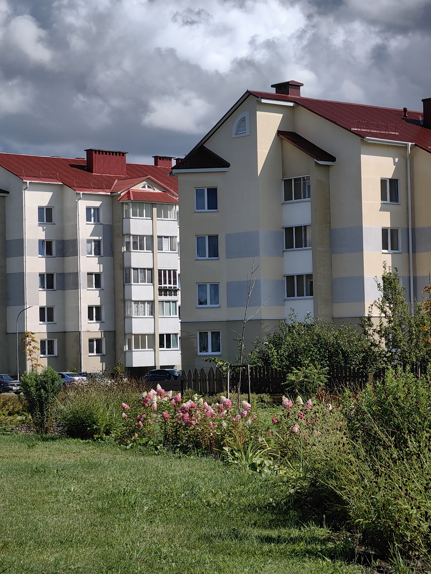 The image shows a residential area in Myadzyel, Belarus. The scene features two apartment buildings with red roofs and beige and gray facades. The buildings have numerous windows, some of which are balconies. In the foreground, there is a well-maintained lawn with a garden bed containing pink and white flowers and various green shrubs. A wooden fence runs along the back of the garden. Several cars are parked in front of the buildings. The sky is partly cloudy, with dark storm clouds visible. The image appears to be taken on a sunny day, possibly late afternoon, given the shadows.