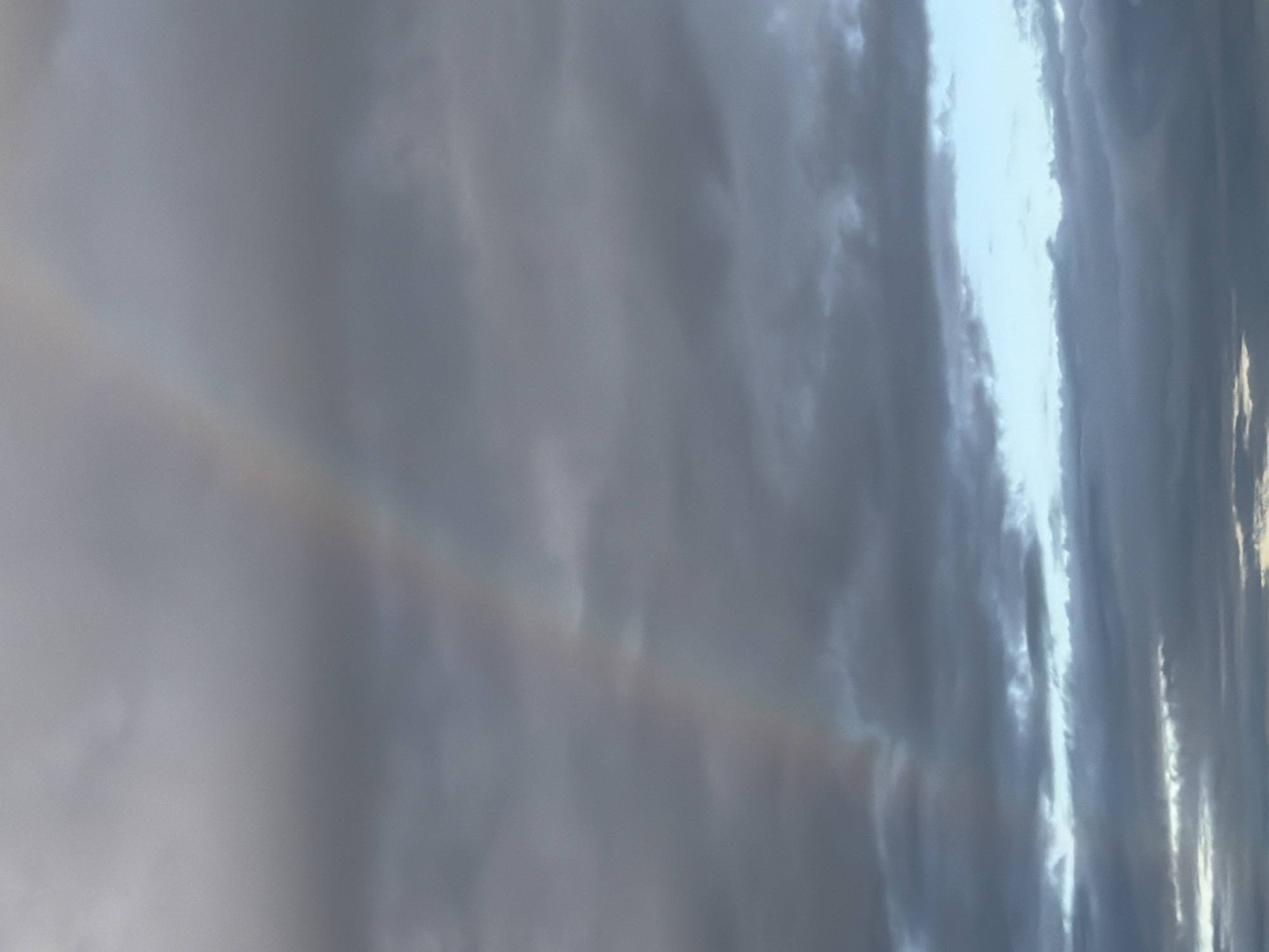 The image displays a view of the sky dominated by gray clouds with a narrow band of lighter, possibly blue, sky visible in the right side. A faint, almost imperceptible rainbow arches across the left side of the image. The location is unknown.