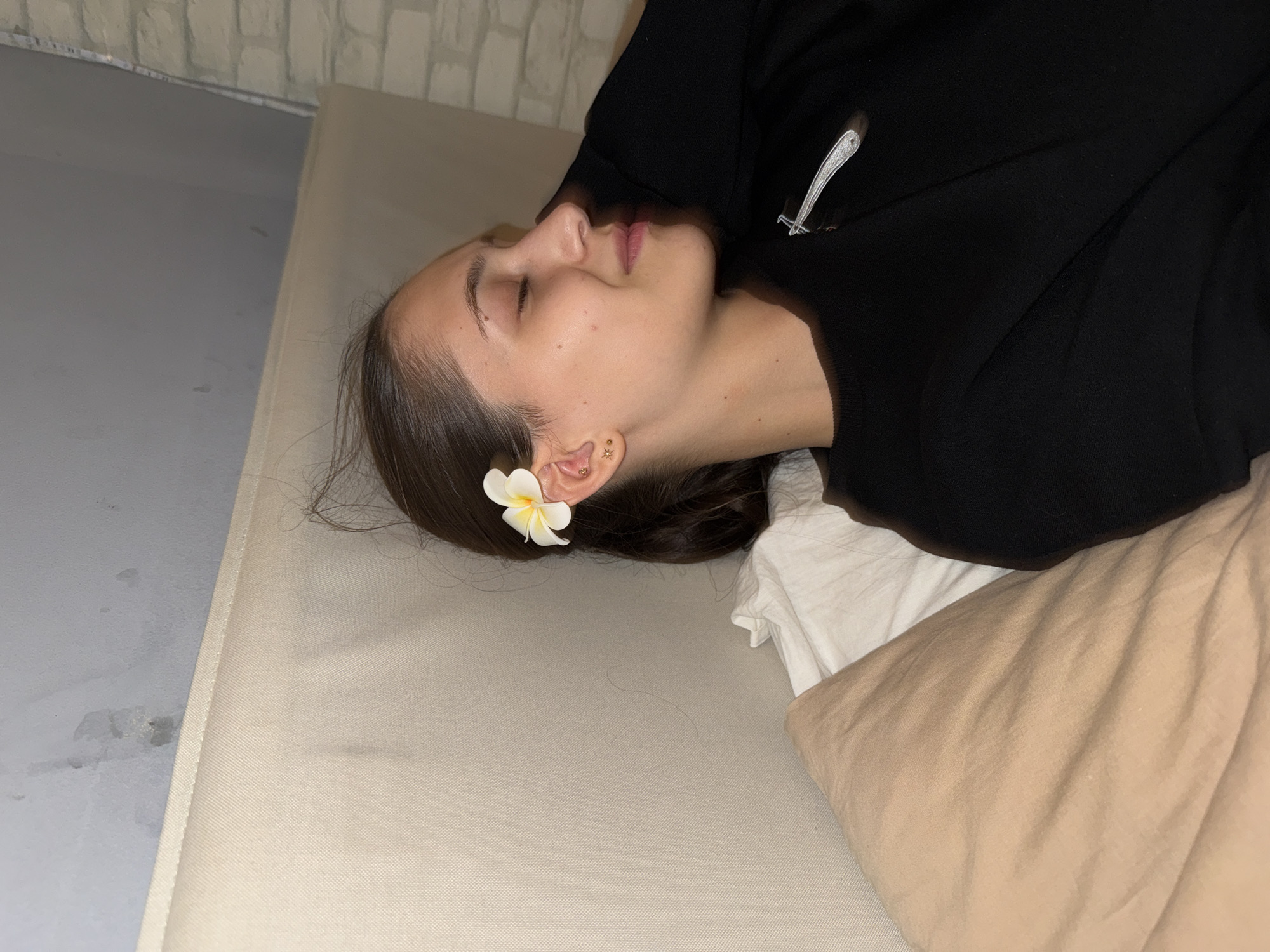 A young woman with her eyes closed and a white flower tucked behind her ear is lying on a bed. She is wearing a black sweater with a silver decorative pin on the chest. Pillows are visible around her. The background shows a textured wall, part of which is light blue and the other part is white brick. This scene is in a bedroom in Sofia, Bulgaria.