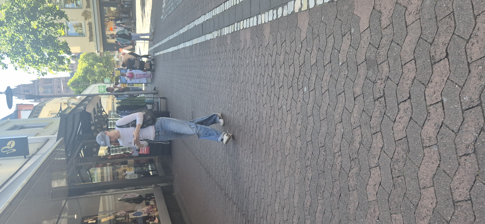 A woman wearing a light-colored long-sleeved shirt, blue jeans, a grey baseball cap with a "NY" logo, and white sneakers walks on a red brick sidewalk in Mainz, Germany. She is holding a mobile phone in her hands and looking down at it, with earbuds in her ears. She carries a black bag on her right shoulder. The street is lined with shops on the left, one with a sign featuring a coffee bean logo. On the right, a street with white and grey paving stones is visible, with other pedestrians in the background, including a person pushing a stroller and a child in pink pants. A tall clock tower with a red-brown spire is visible in the distance, peeking above the buildings and trees, likely the Mainz Cathedral given the location. The sun is shining, casting shadows.