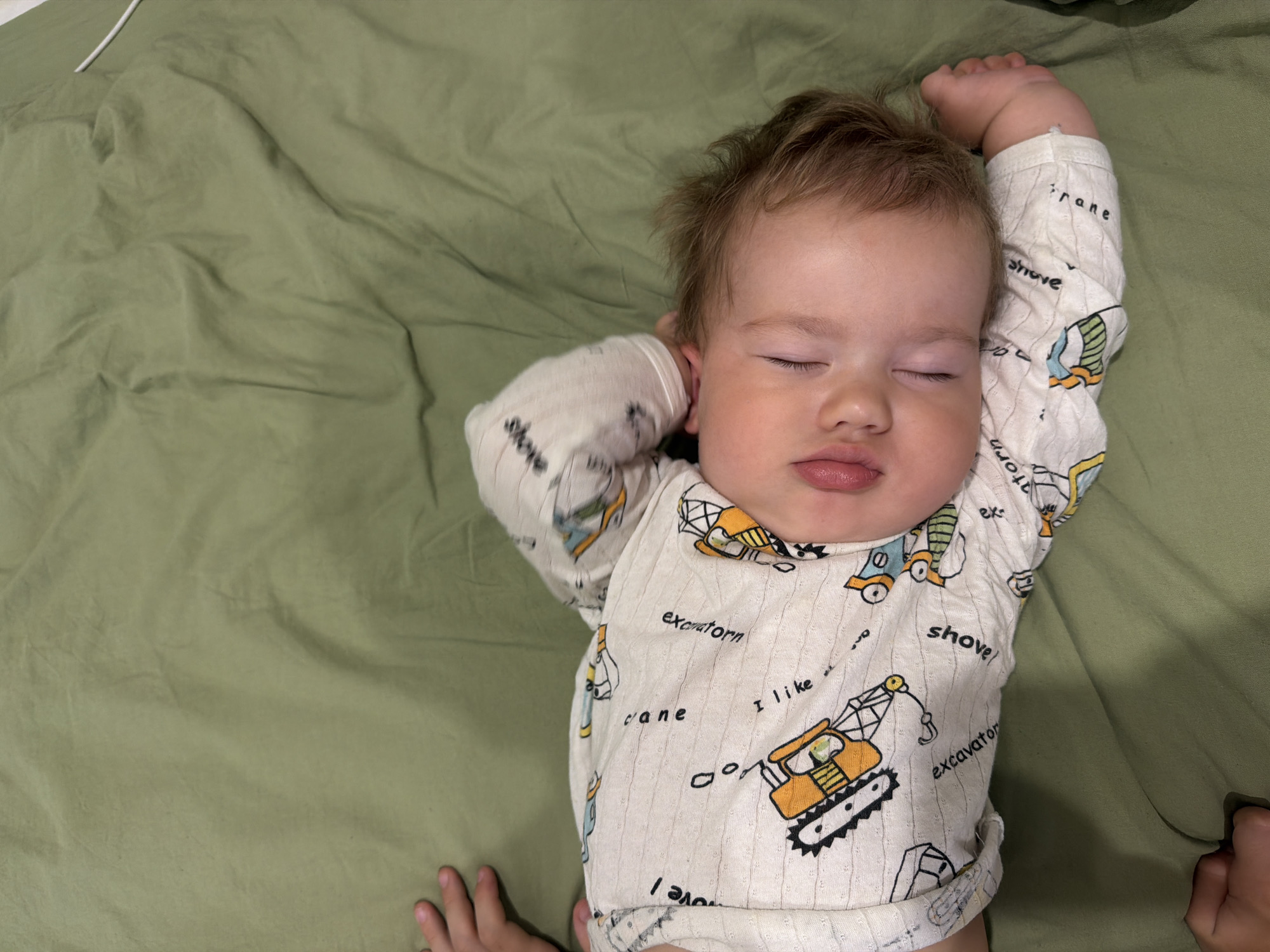 A baby is sleeping on a green fabric surface. The baby is lying on its back, facing upwards. The baby is wearing a white pajama set with images of construction vehicles and text. The baby's mouth is slightly open, and its tongue is visible. The baby's right arm is bent and resting on its head, while its left arm is bent at the elbow. The baby's feet are visible at the bottom right of the image. The green fabric appears to be a bedsheet or blanket, and it is wrinkled in places, suggesting it is in a domestic setting, possibly a bedroom. The lighting in the image is soft, indicating it might be taken during the day with natural light or with artificial lighting that is not harsh. There is no discernible information in the image to determine its location in Kazan, Russia.