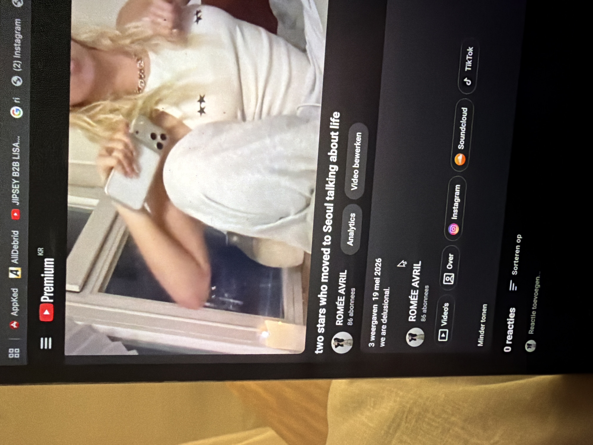 The image shows a screen capture of a YouTube video interface. The video is titled "two stars who moved to Seoul talking about life" by a channel called ROMÉE AVRIL with 86 subscribers. The video was uploaded on May 19, 2026, and has 3 views. The description of the video is "we are delusional." On the left side of the screen, app icons for AppKed, AllDebrid, YouTube Premium, JIPSEY B2B LISA, Instagram, and Google are visible. On the right side, options for Instagram, Soundcloud, and TikTok are shown. Below the video, there is information about the channel, including the channel's name, subscriber count, video list, and the "Over" tab. Below that, there are options to hide sounds, and the option to sort the video. No reactions are seen, and there is a field to add a reaction. The image itself shows a person with blonde hair holding a smartphone or similar device. The person is wearing a white shirt with star designs. The background appears to be an indoor setting with a window. There is no further information about the city of Seoul, South Korea.