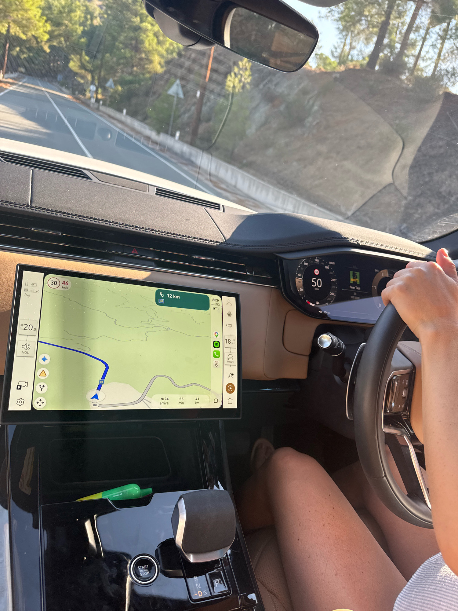 An outdoor, high-angle, full shot captures the interior of a modern vehicle as it drives on a paved road through a wooded area. Sunlight illuminates the scene, casting shadows on the road and trees. The driver's hands are visible on the steering wheel, and their legs are also in view, suggesting they are wearing a dress or skirt. The car's dashboard features a large touchscreen display showing a navigation map with a route highlighted in blue, indicating a journey of 12 km. The digital speedometer displays a speed of 50 km/h. The vehicle's central console contains the gear shifter and a start/stop engine button. A green pen lies on the console. The environment outside the car consists of a road, trees, and vegetation, with the bright sky visible above. The scene is set in Moniatis, Cyprus, as indicated by the navigation system.